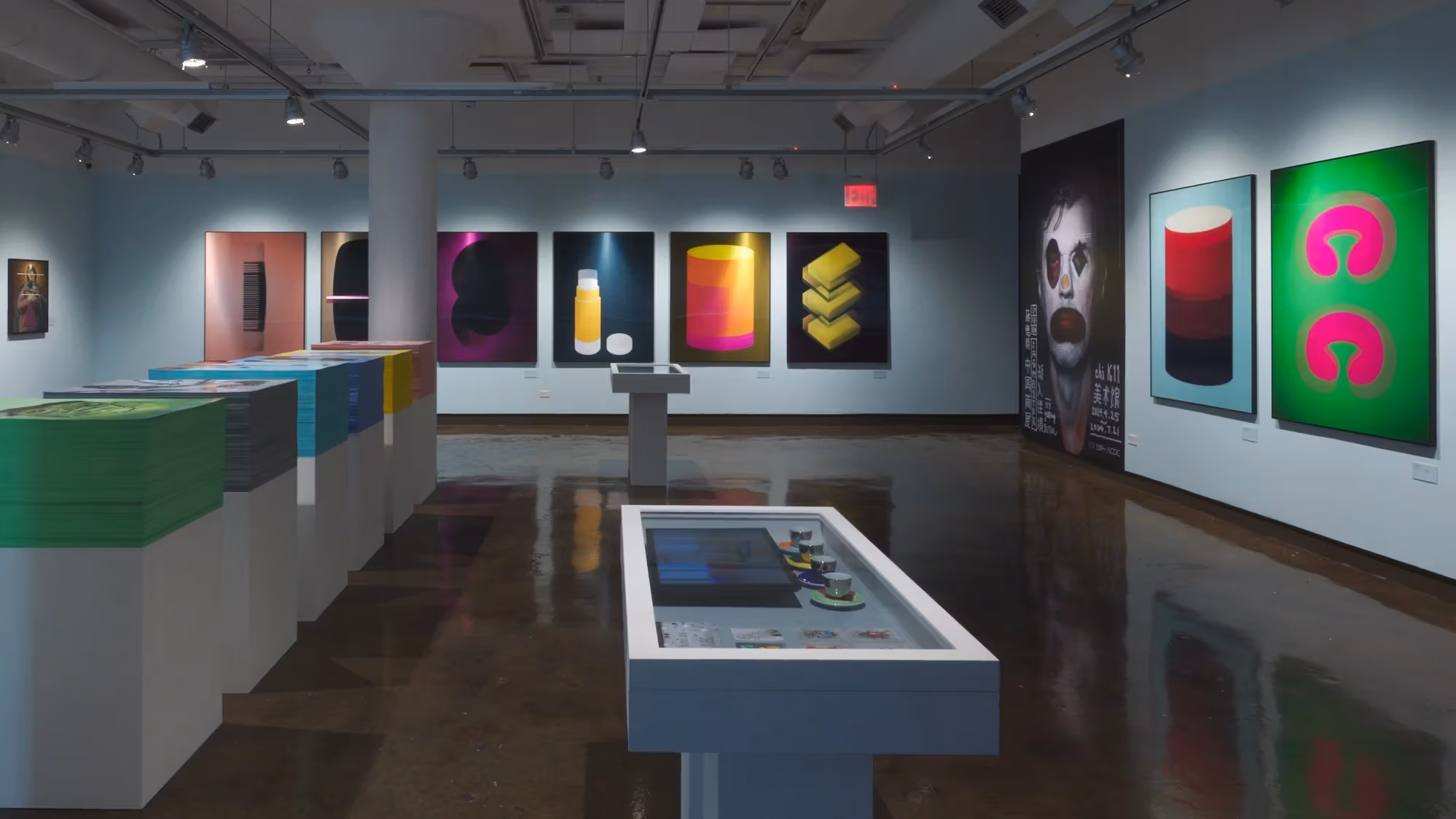 Wide view of an art exhibition in situ at SVA Chelsea Gallery including work hung on walls, vitrines and large stacks of ephemera for visitors to take.