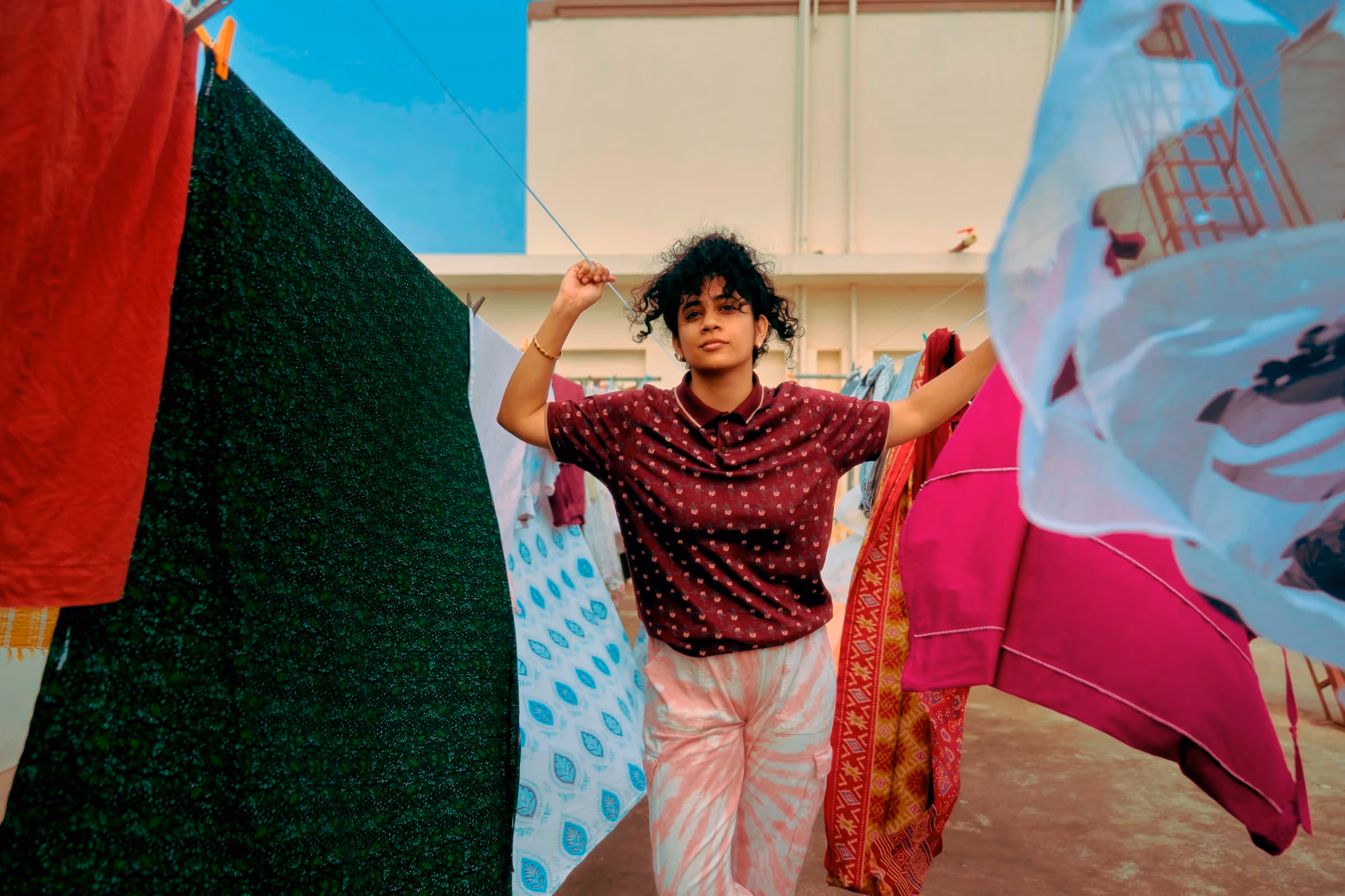 In this color self-portrait, the artist Shravya Kag is standing among pieces of fabric drying on cloth lines. She is wearing a short sleeve top in burgundy red with yellow polka dots, and a pair of tie-dyed long pants in salmon pink and white.