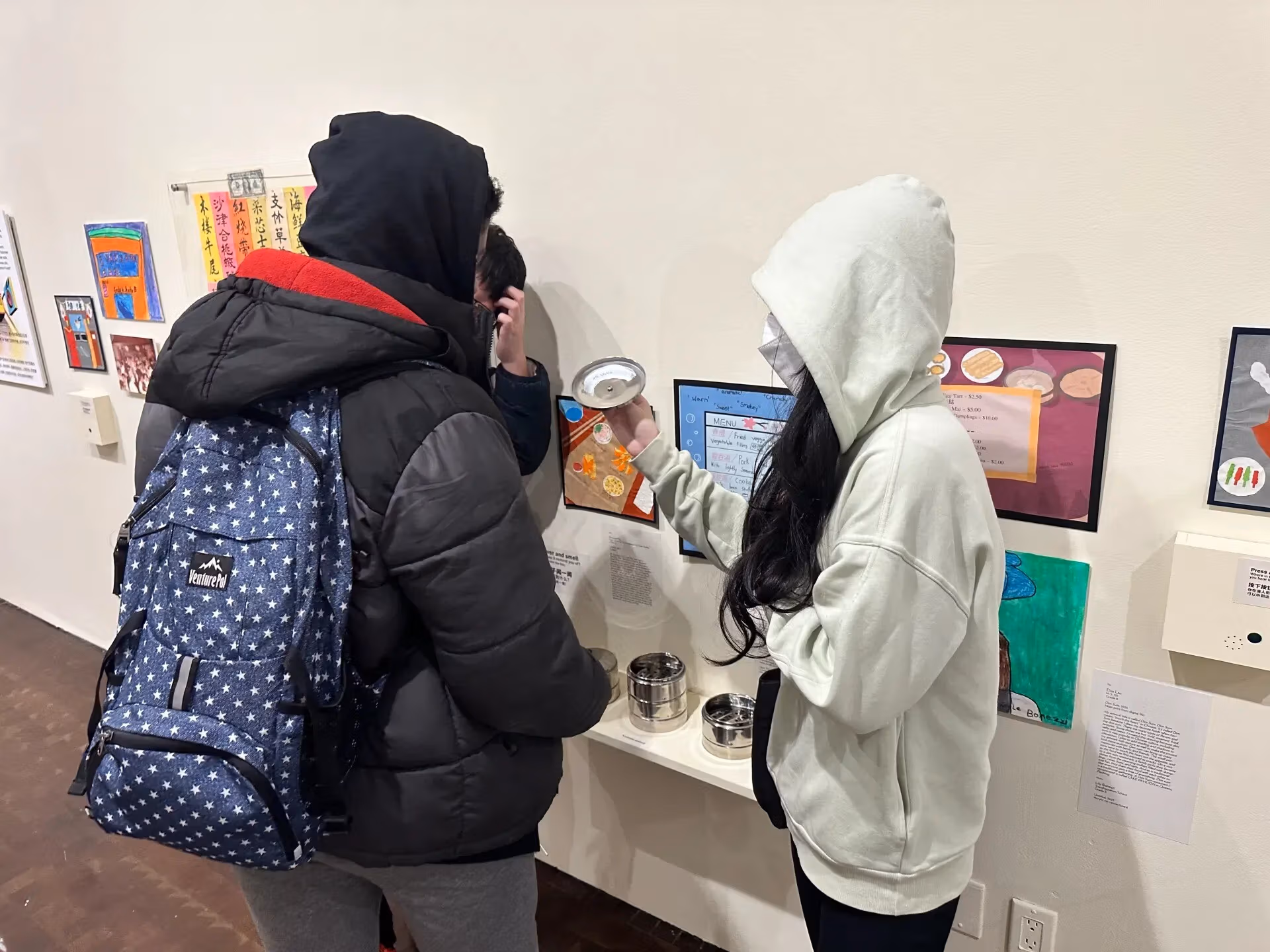 Picture of a few young people in a museum exhibition, inspecting one of the artworks that seems to involve a lid, which one of them is holding, and some containers on a small shelf on the wall.