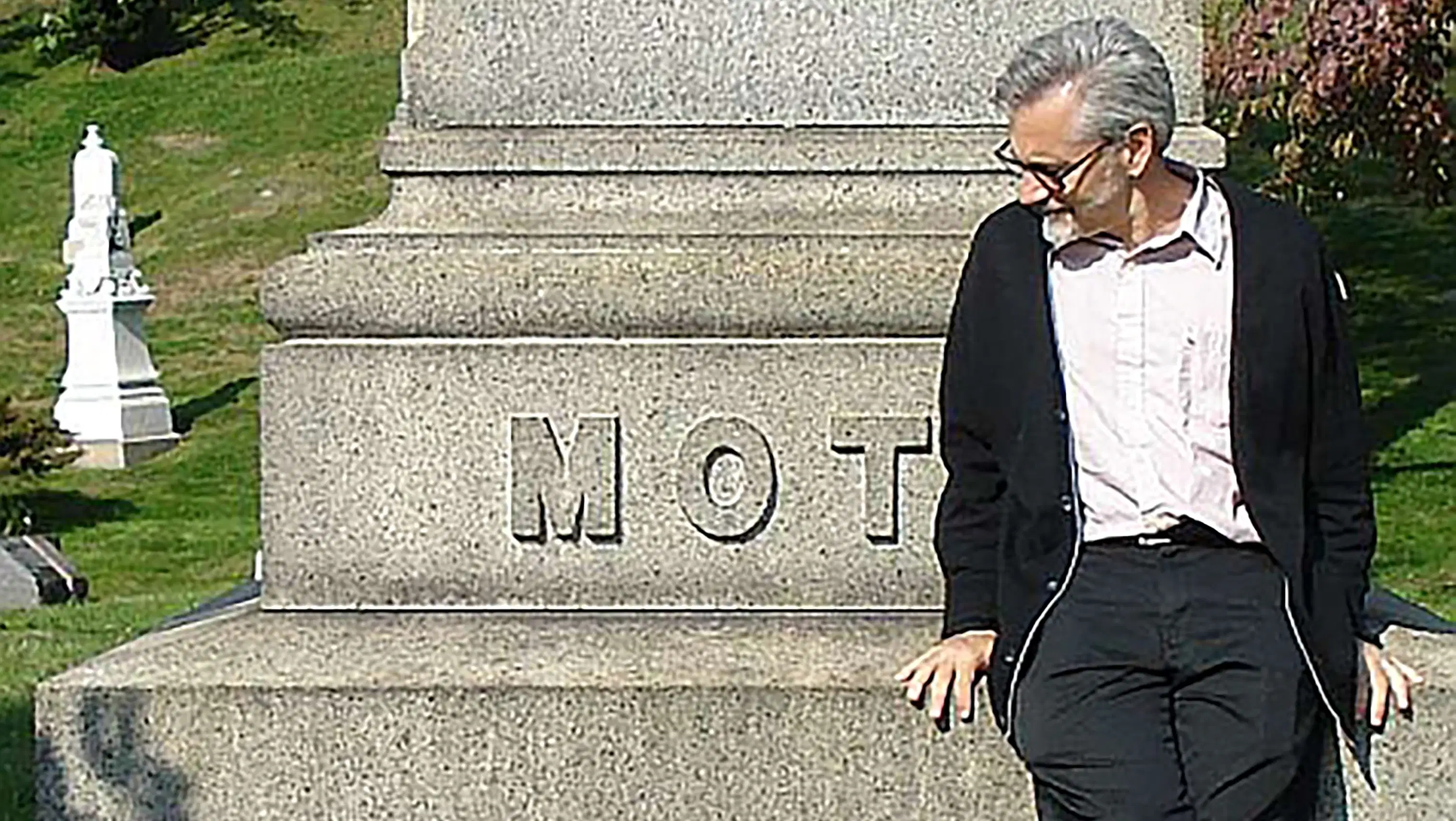 Cartoonist tom motley in a graveyard sitting on a large memorial gravestone that says "MOT"
