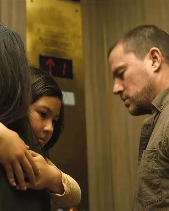 A person holds a child protectively in an elevator, while another person stands nearby, looking down. The atmosphere is tense and somber, with muted tones.