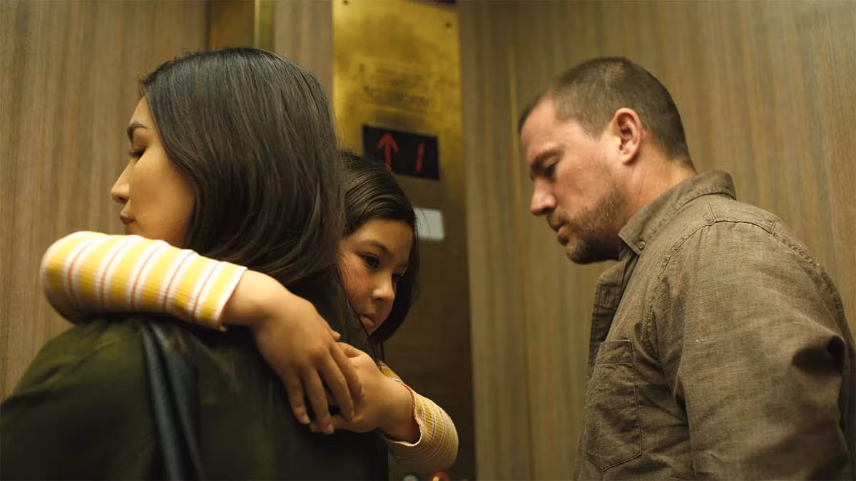 A person holds a child protectively in an elevator, while another person stands nearby, looking down. The atmosphere is tense and somber, with muted tones.