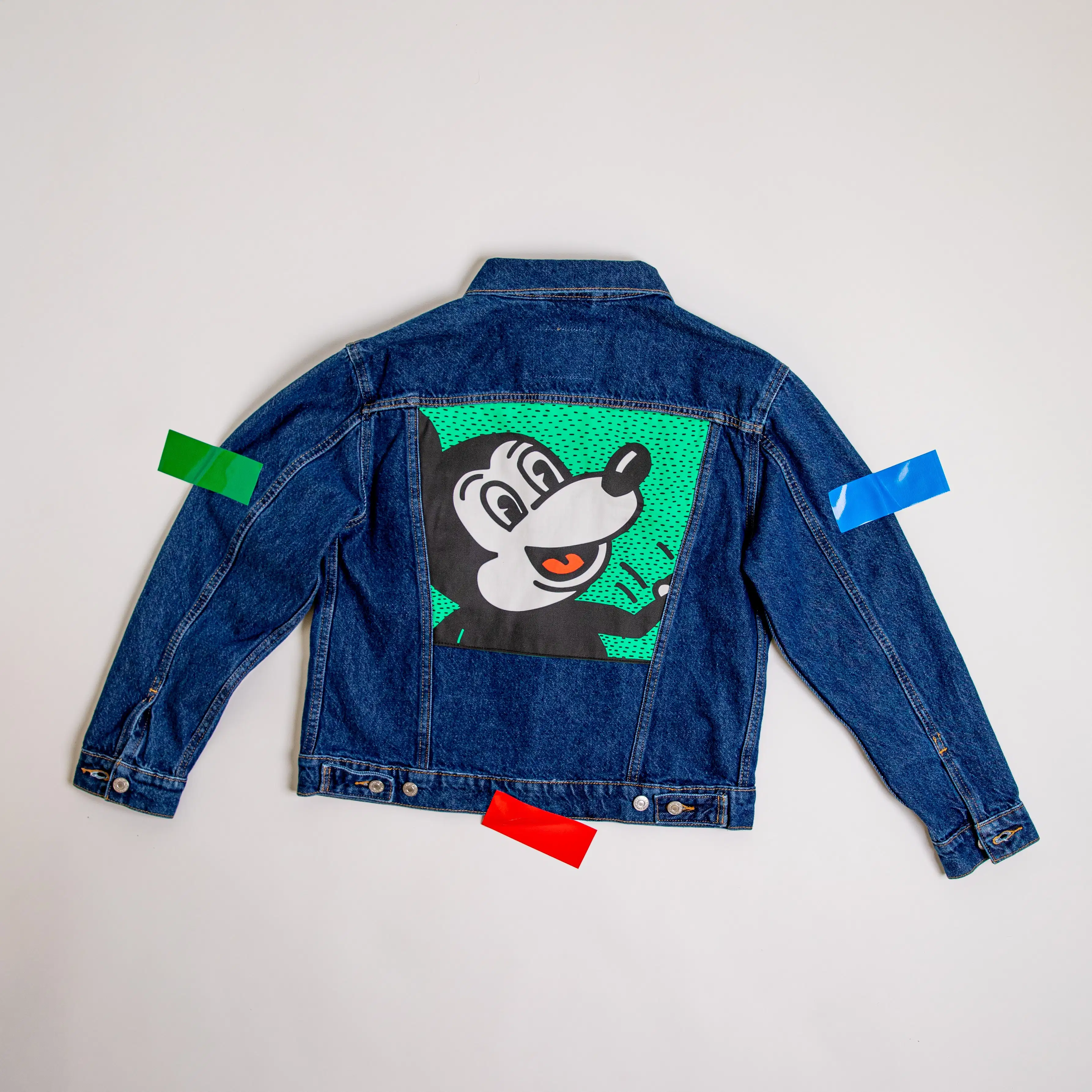 A photograph of a jean jacket with a Keith Haring illustration of Mickey Mouse on the back. The jacket is taped to the surface it is on.