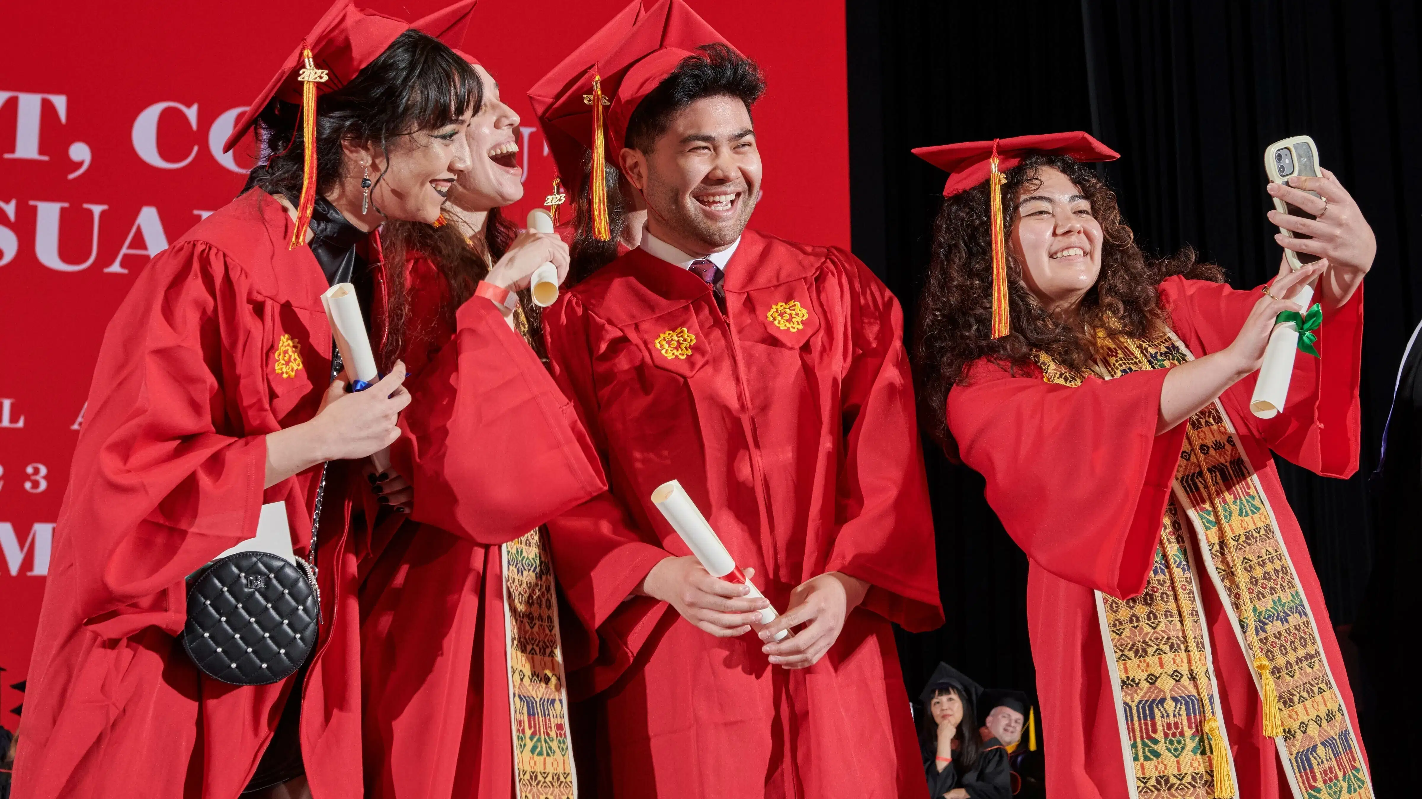 SVA graduating students in red caps and gowns, some holding flowers, smiling and posing for photos inside Radio City Music Hall in New York.