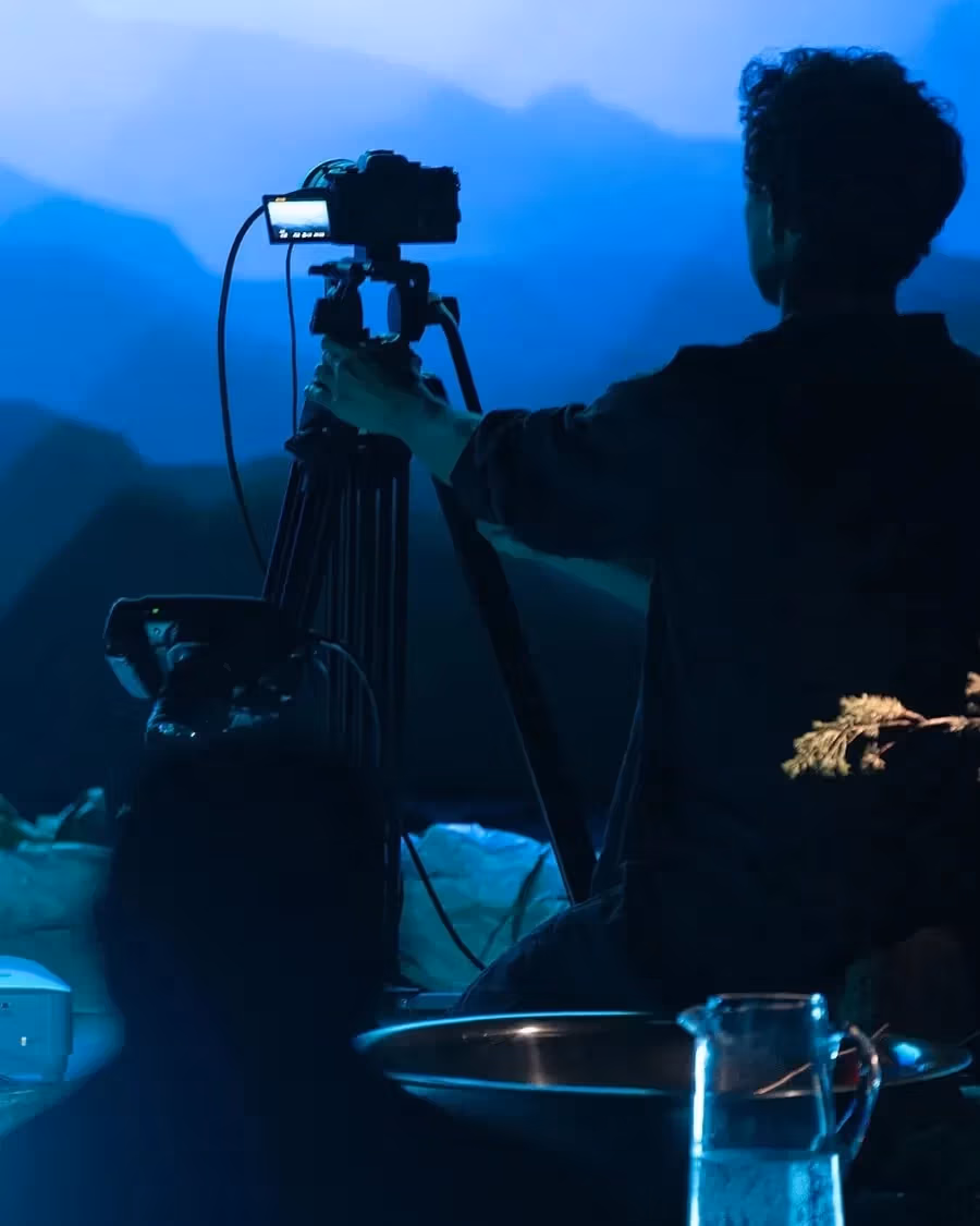  Image of a person pointing a recording camera on a tripod towards a wall with a projected image of what looks like a mountain range.