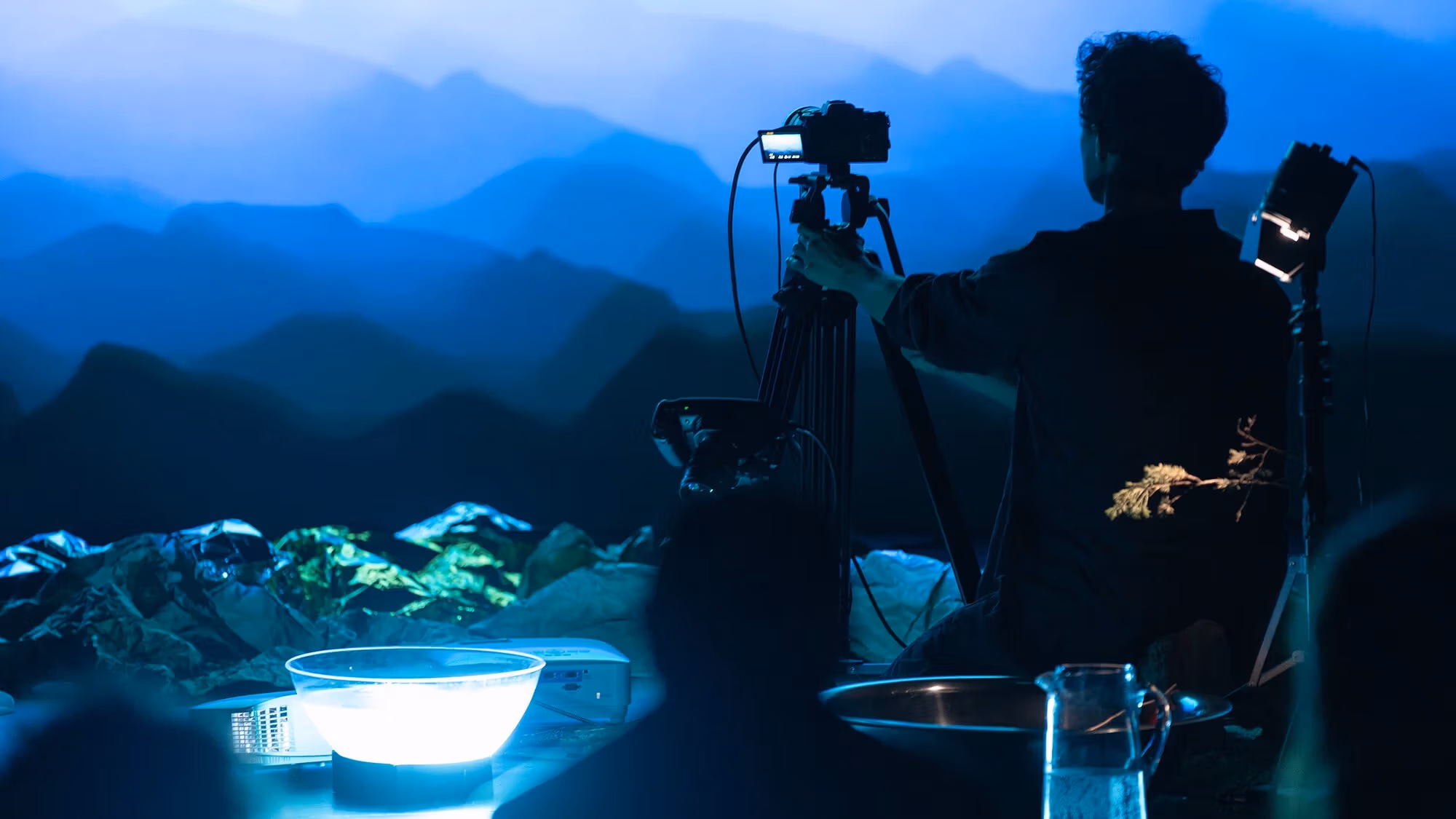  Image of a person pointing a recording camera on a tripod towards a wall with a projected image of what looks like a mountain range.