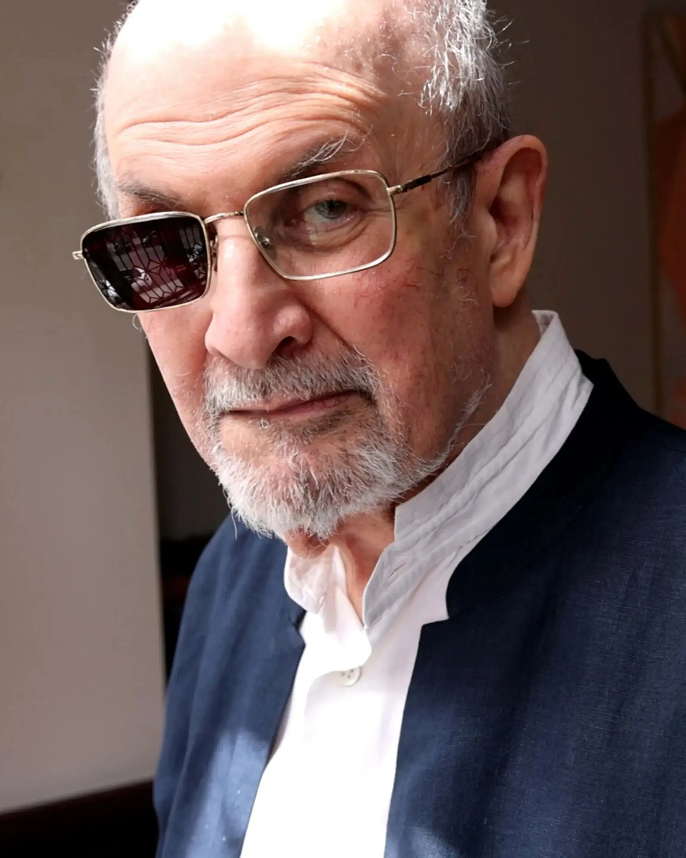 A person with a serious expression is indoors, wearing a dark jacket over a white shirt. One lens of his glasses is shaded. Soft lighting adds a contemplative tone.