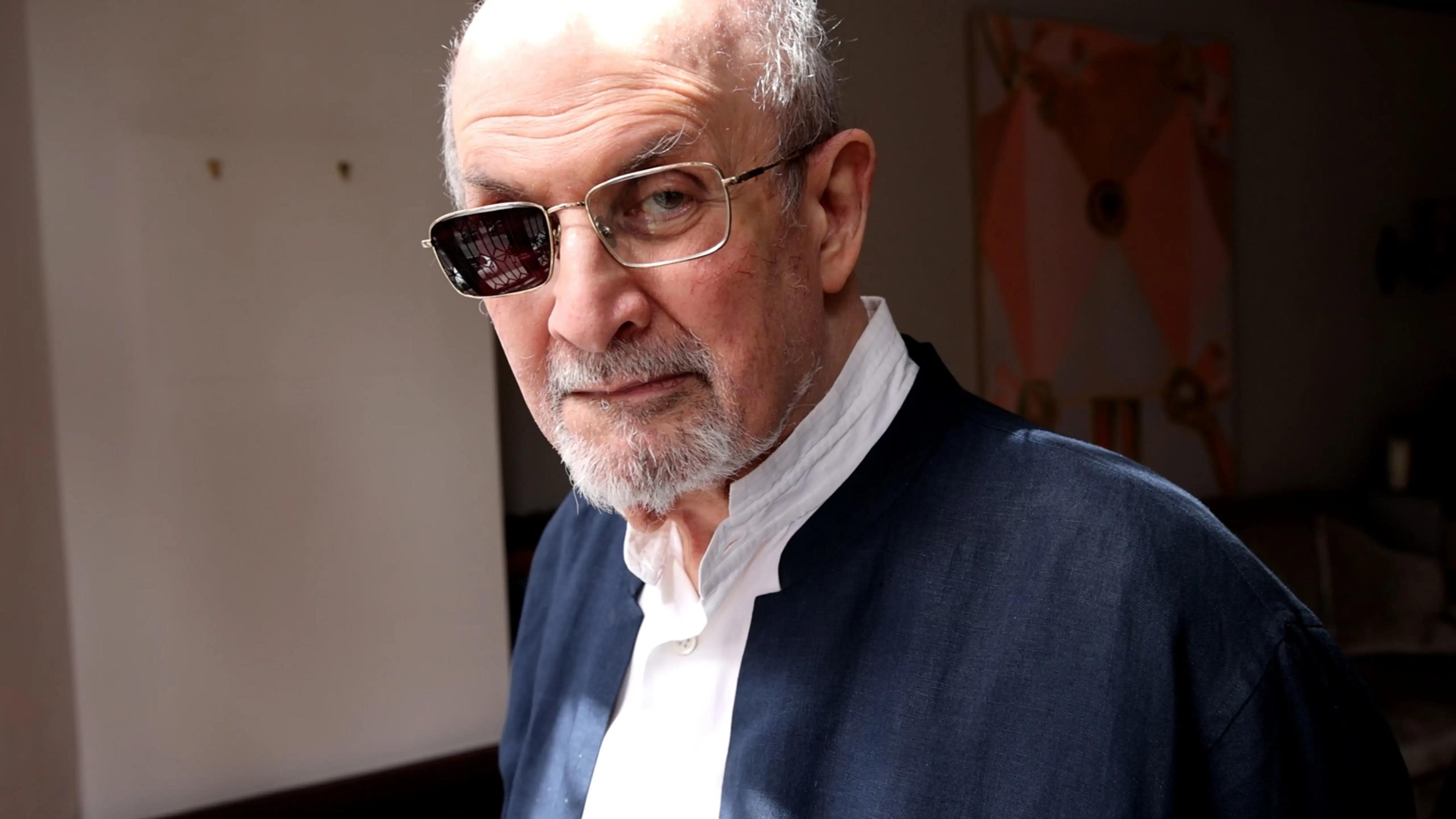 A person with a serious expression is indoors, wearing a dark jacket over a white shirt. One lens of his glasses is shaded. Soft lighting adds a contemplative tone.
