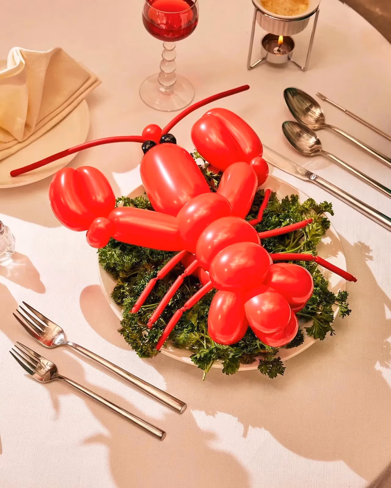 A photograph of a lobster shaped red balloon sitting on top of a bed of kale leaves on a dinner plate. Surrounding the scene is a dinner table fully set up with cutlery,  a dinner napkin folded into a fan, and a single rose in a tin vase.