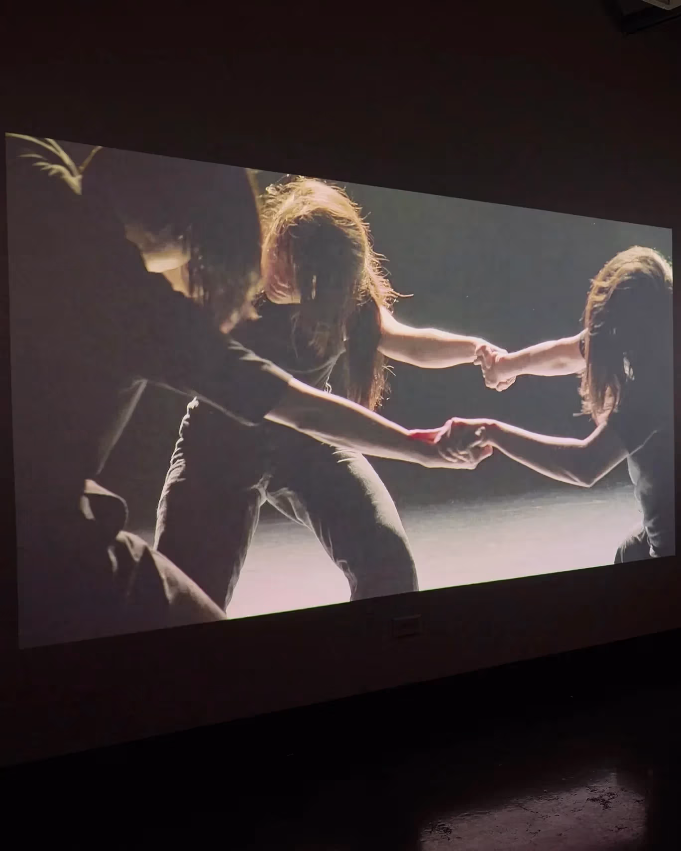 An installation view of two lens based artworks in a dark room. The one on the left is an image of three people holding hand. The image on the right is a still of a woman documenting a performance.