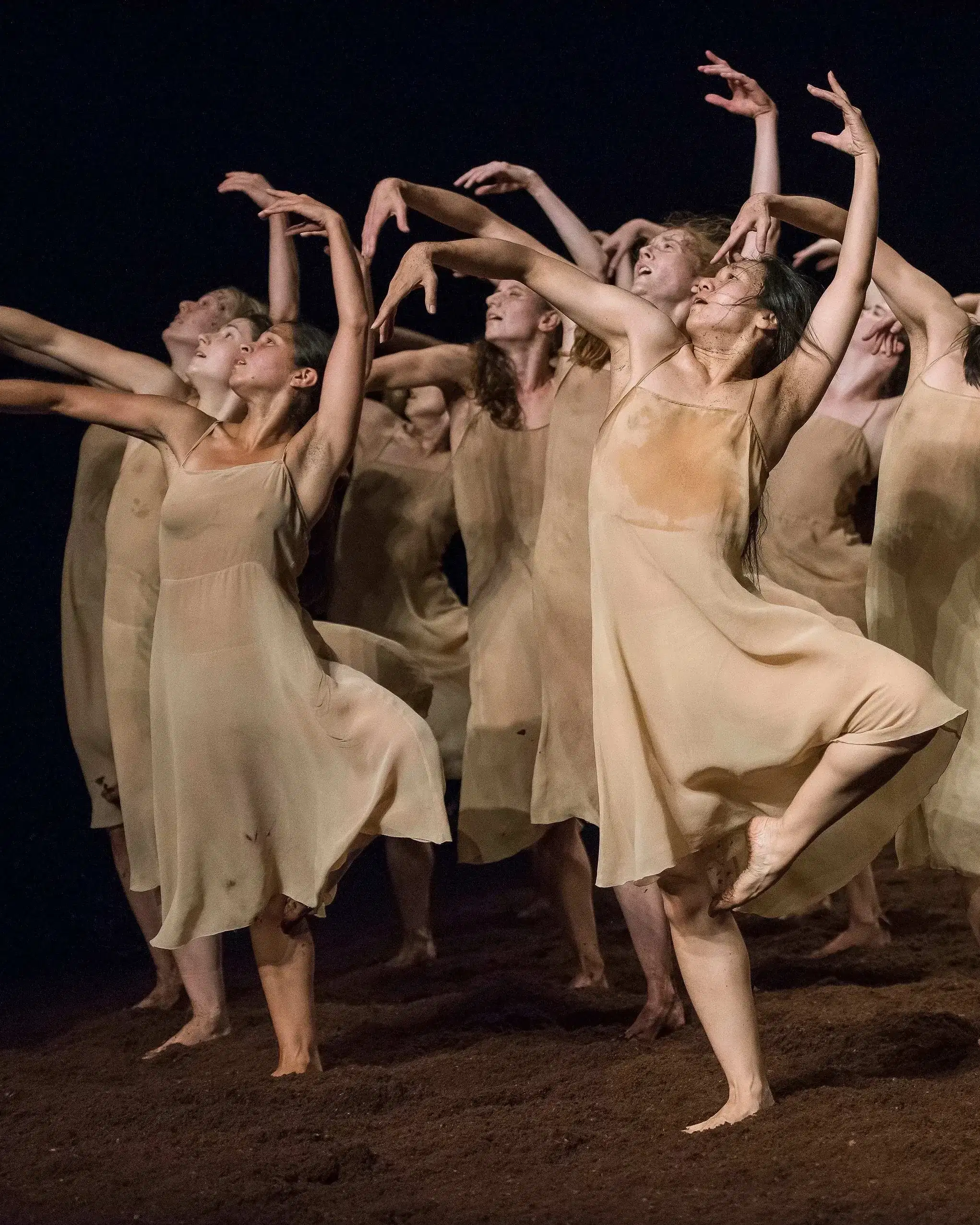 A group of dancers in beige dresses on a dirt floor standing on one leg with the other leg bent, holding their hand above their heads while looking up and to the left.