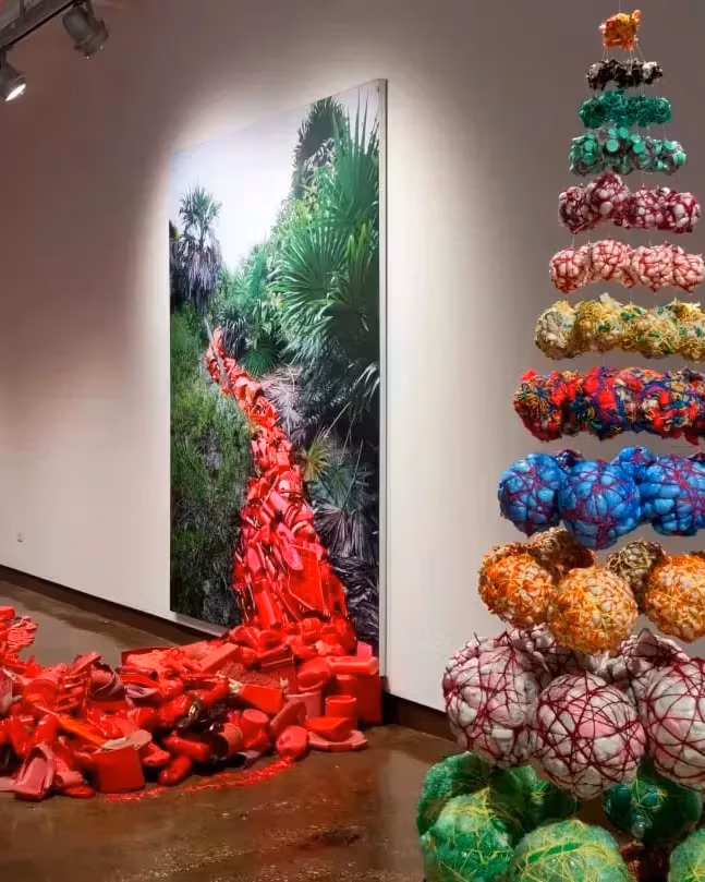 A photograph of an art exhibition featuring two sculptural works—one a collection of red objects that extends from a photograph on the wall, the other a pyramid-shaped arrangement of spherical objects.