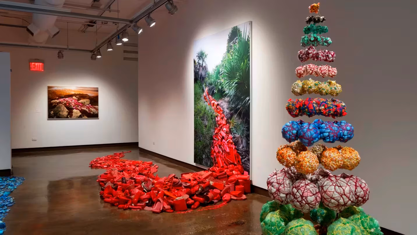 A photograph of an art exhibition featuring two sculptural works—one a collection of red objects that extends from a photograph on the wall, the other a pyramid-shaped arrangement of spherical objects.
