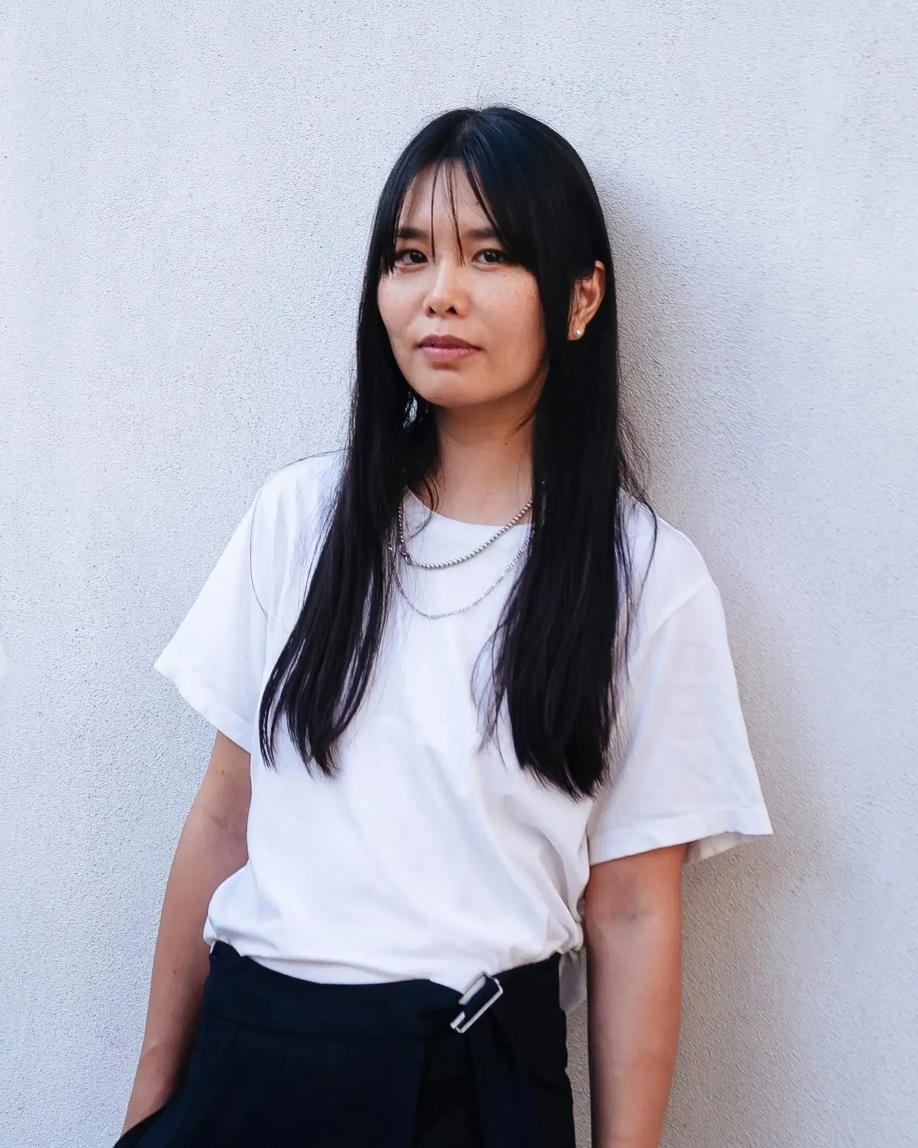 A photo of Ami Lien. She is leaning against a white wall and looking into the camera with a relaxed expression. She is wearing a white shirt and black pants and has two silver chain necklaces on.