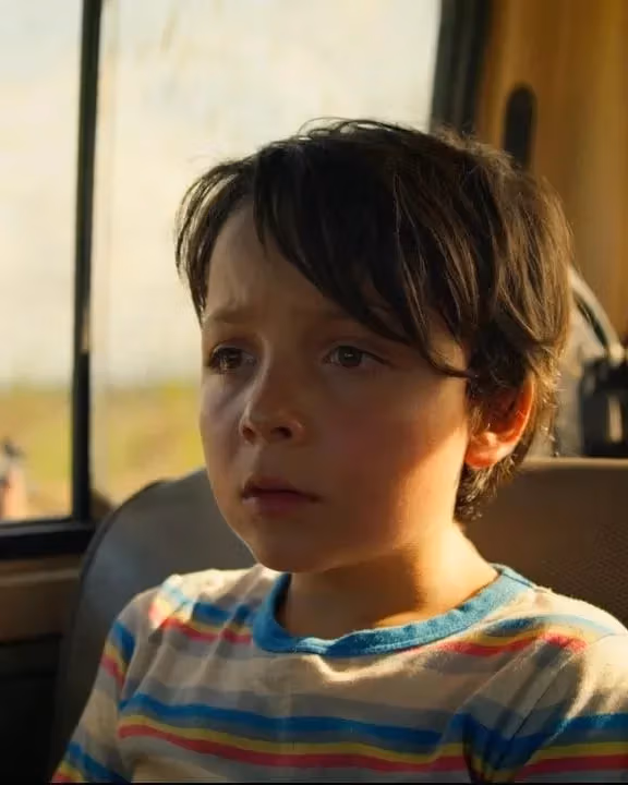  A young boy sitting in the back seat of a vehicle, looking out the window with a thoughtful expression.