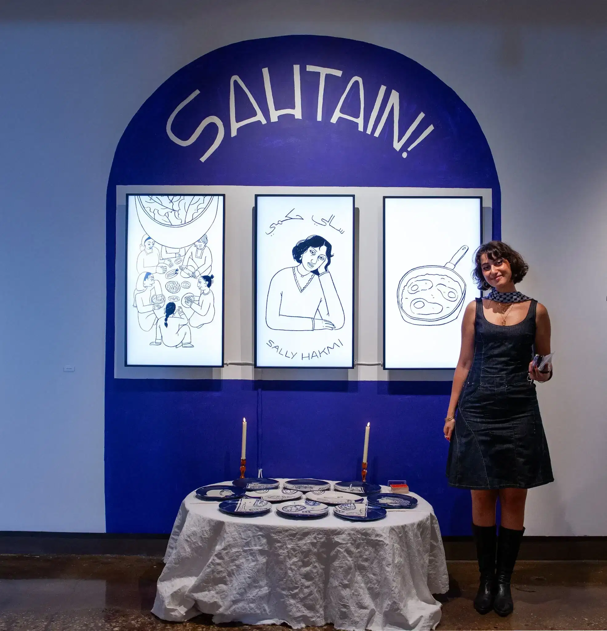 A person stands in front of an exhibit featuring a table dressed with plates and candles in front of a wall with a painted blue arch and three screens showing off illustrations.