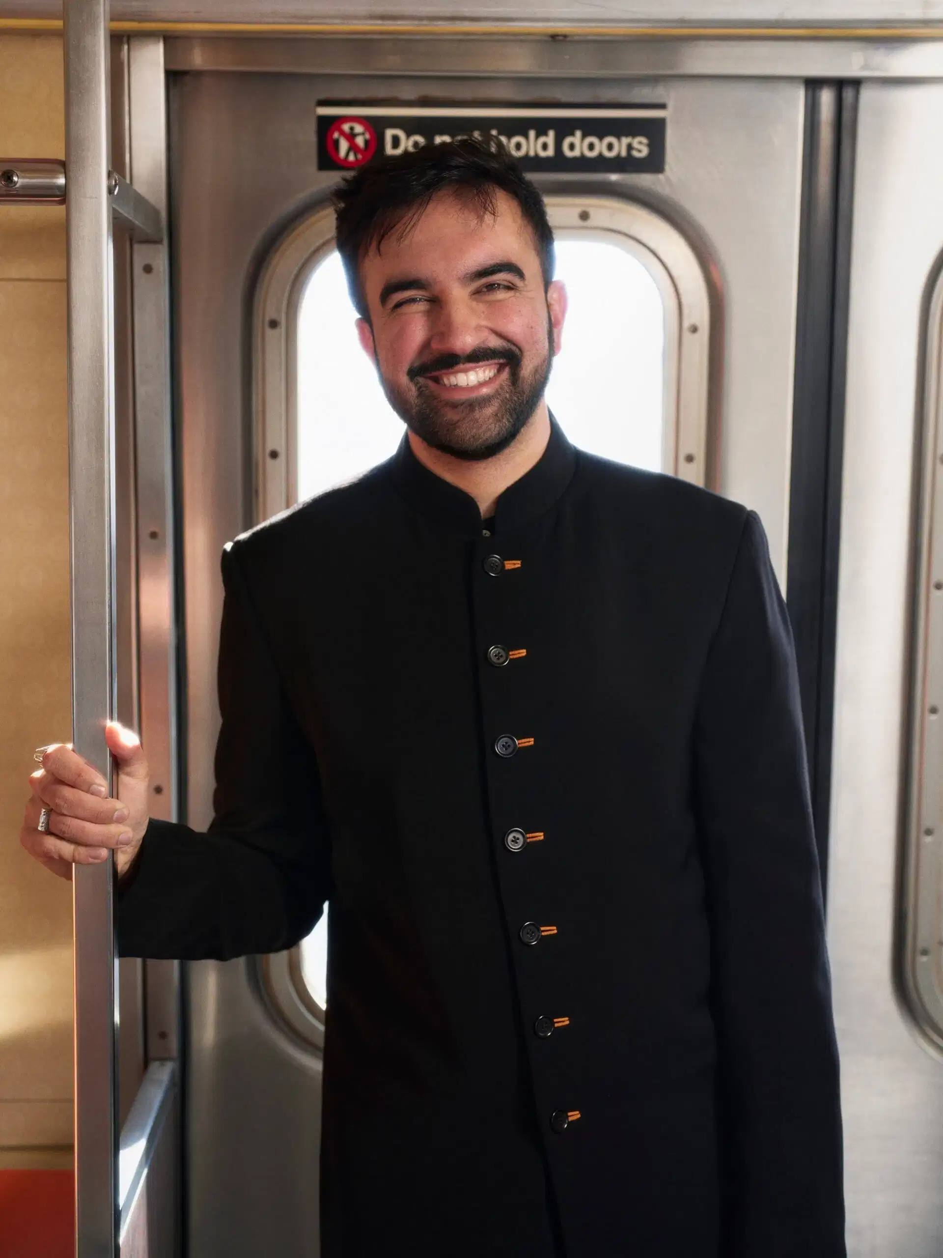 A person stands on the subway in a black button up. They are smiling widely and warmly.