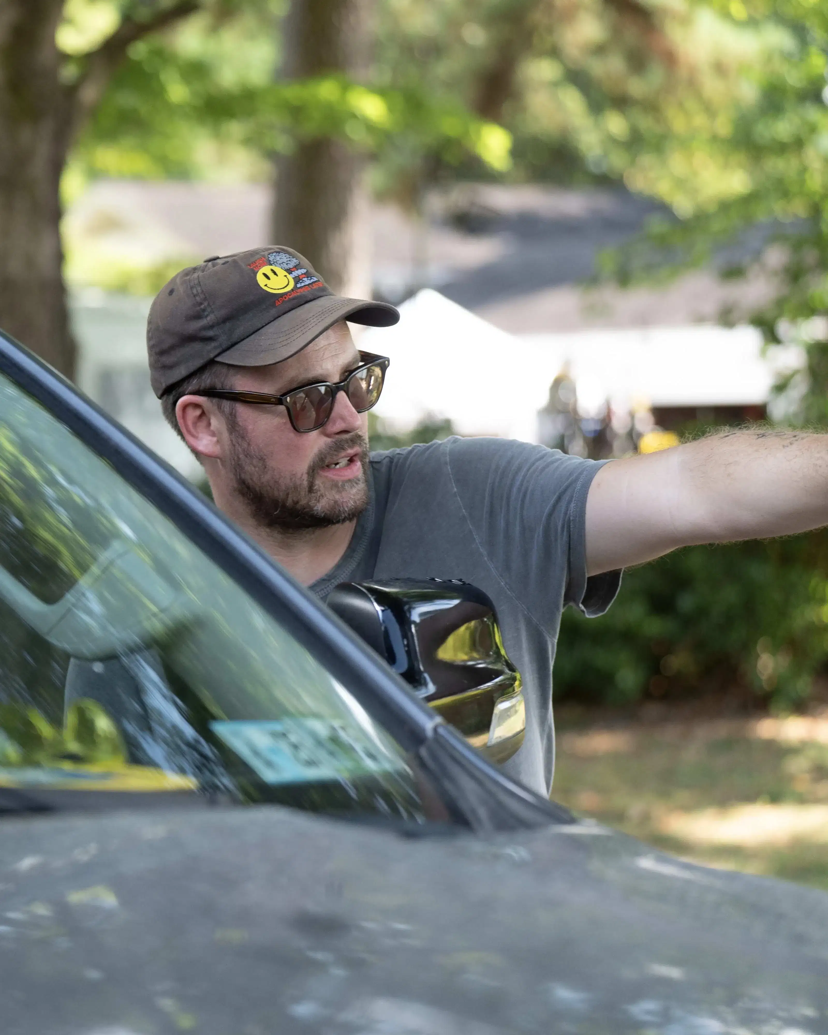 A man sits focused in a car, looking forward. Another man outside gestures and directs, wearing glasses and a cap. Trees create a serene, green backdrop.