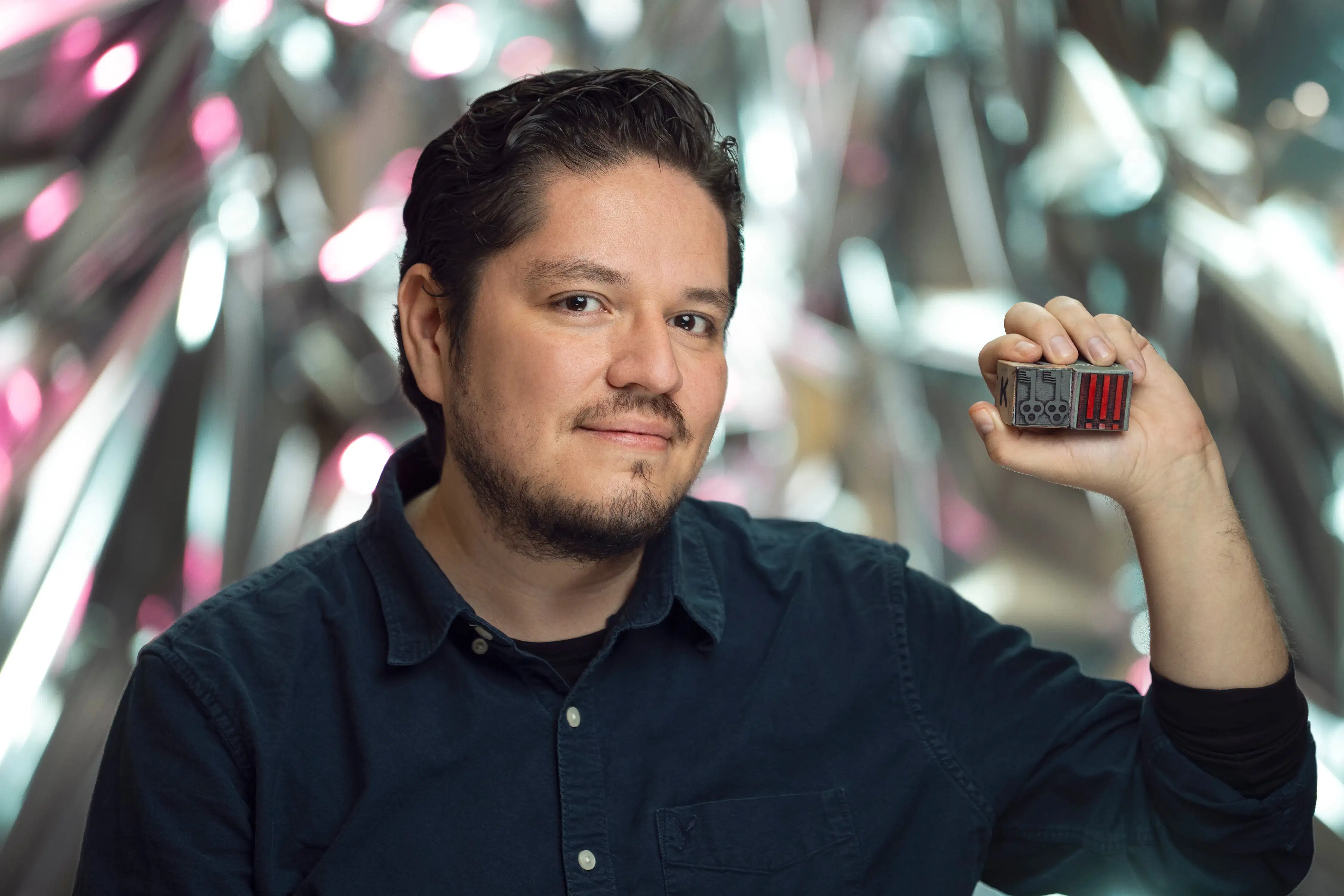 A man in front of a reflective background smiles at the viewer while holding up a small figure in his left hand.