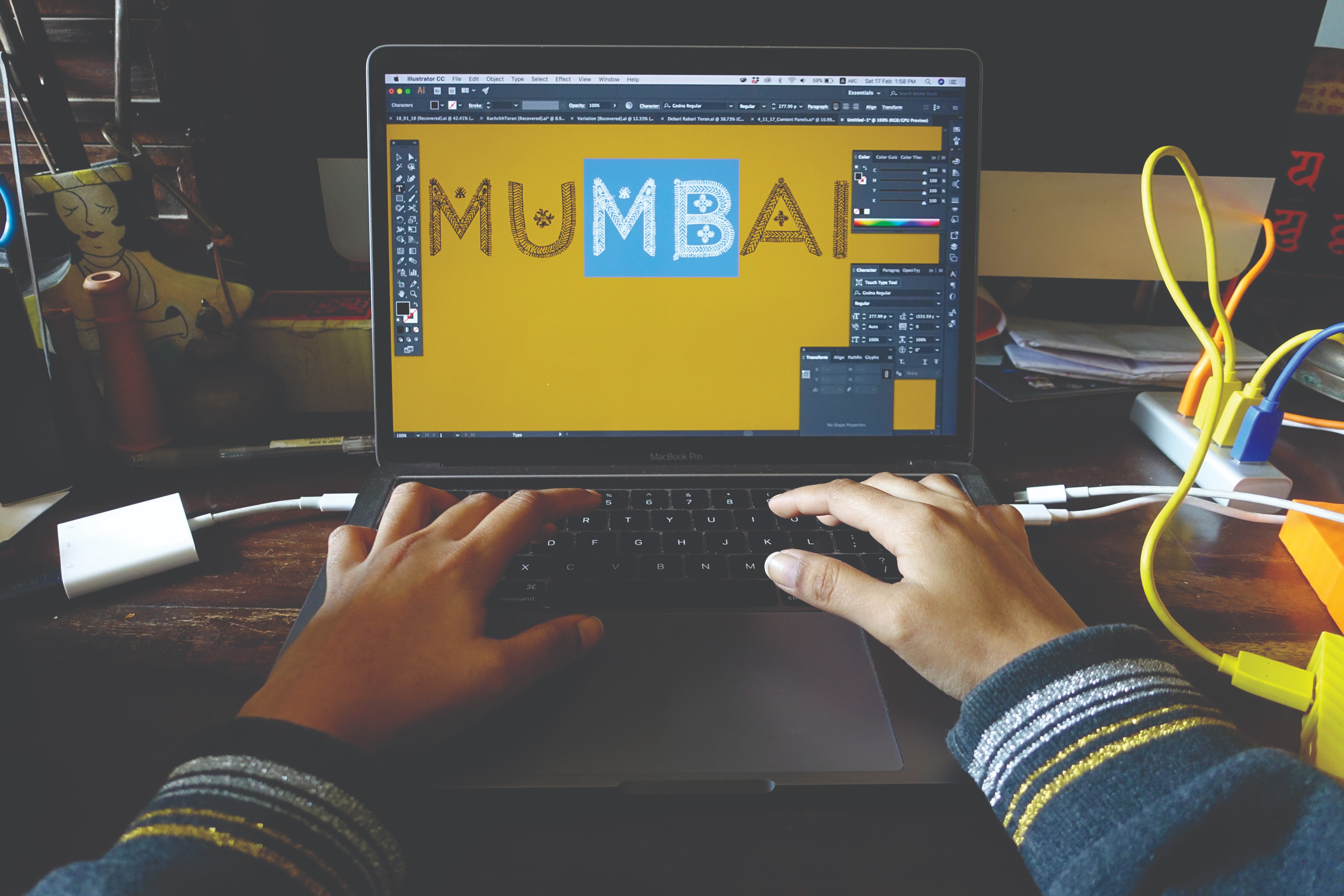 A color photograph of two hands typing on a laptop, which is displaying the text "Mumbai" in all capital letters.