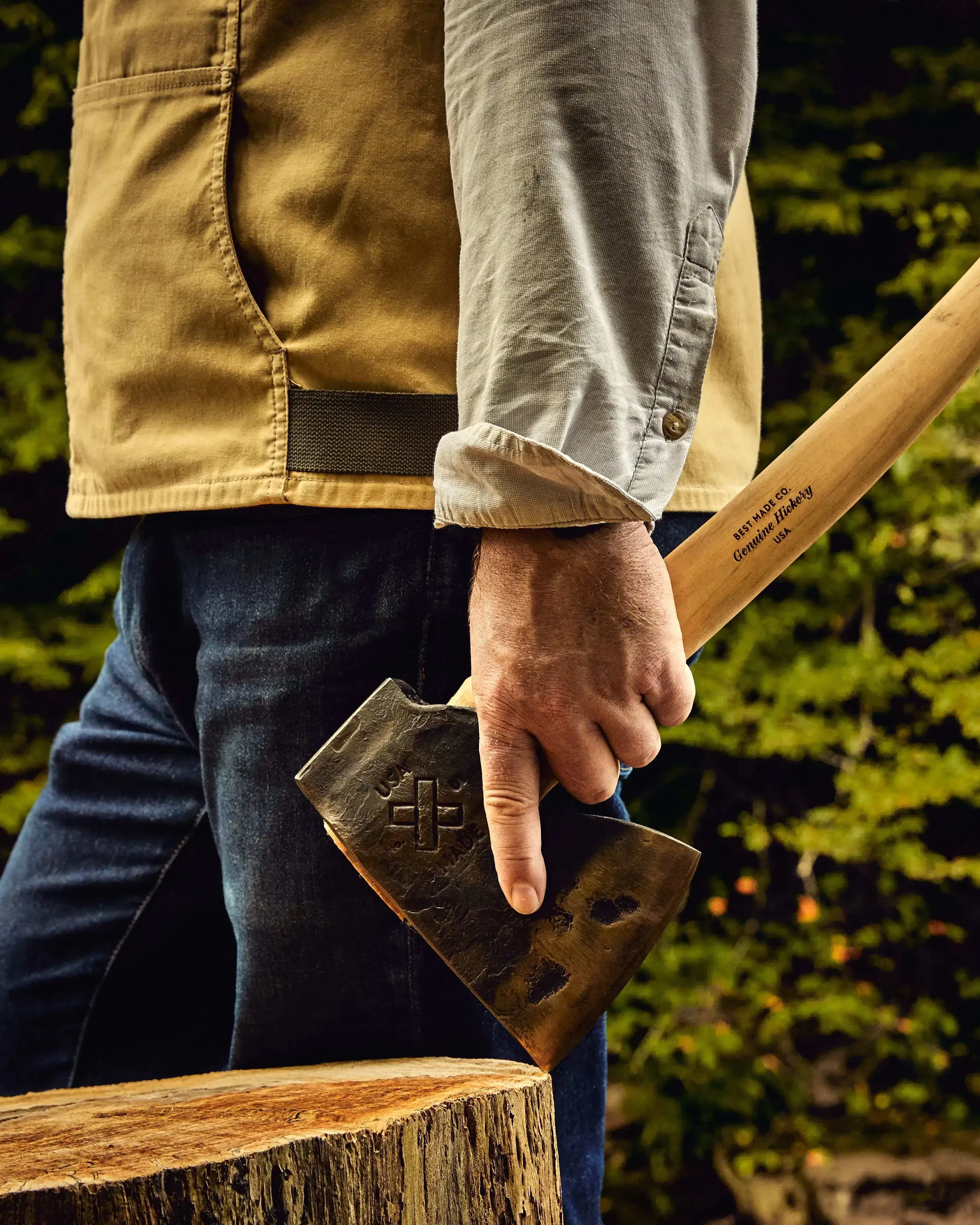 A person standing outdoors holds an axe by their side, with a tree stump in front and trees in the background.