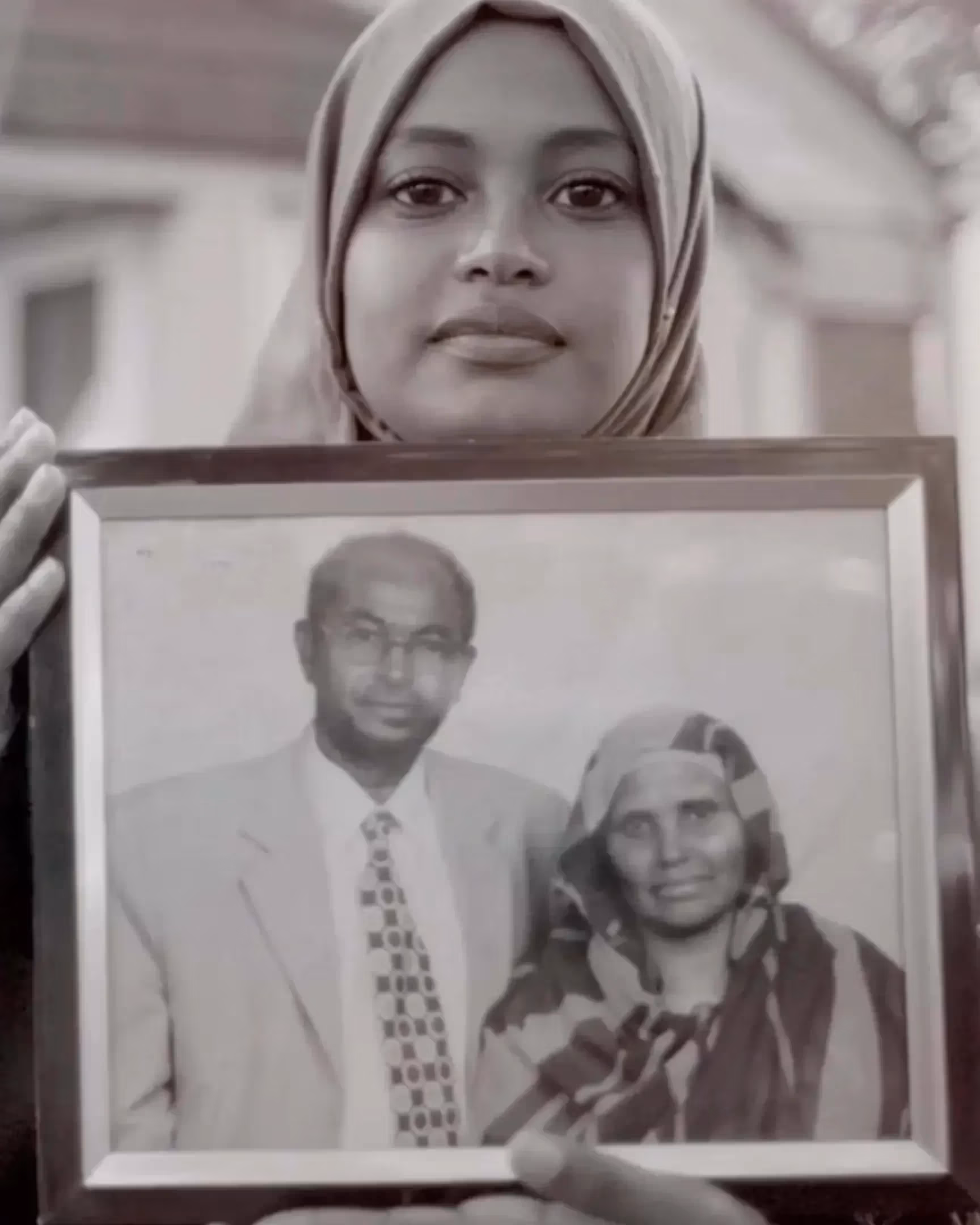 Black and white image of a woman in a hijab holding a picture frame. The picture in the frame is of a man in a suit and another woman in a hijab.