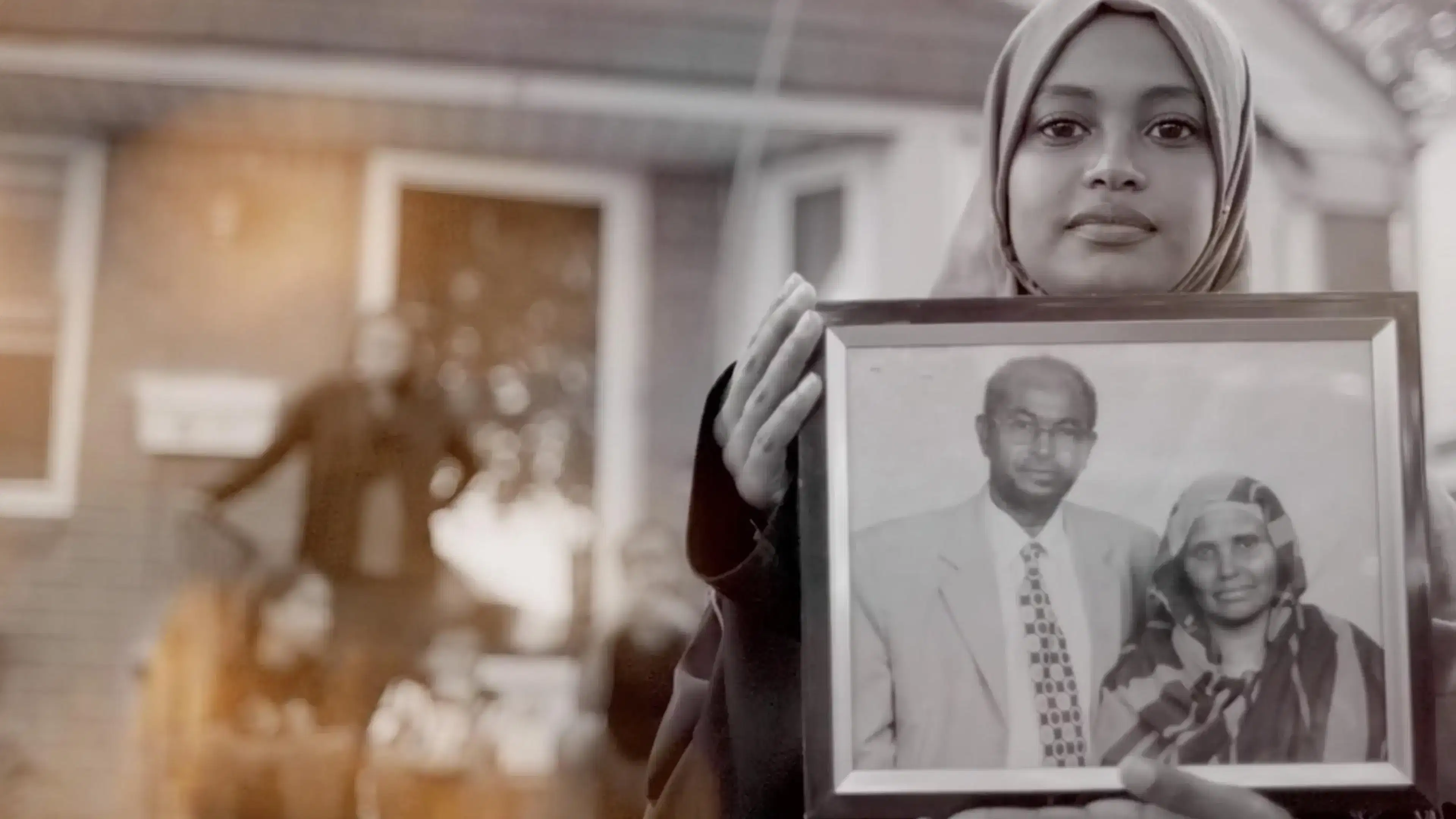 Black and white image of a woman in a hijab holding a picture frame and a man out of focus on the left. The picture in the frame is of a man in a suit and another woman in a hijab.