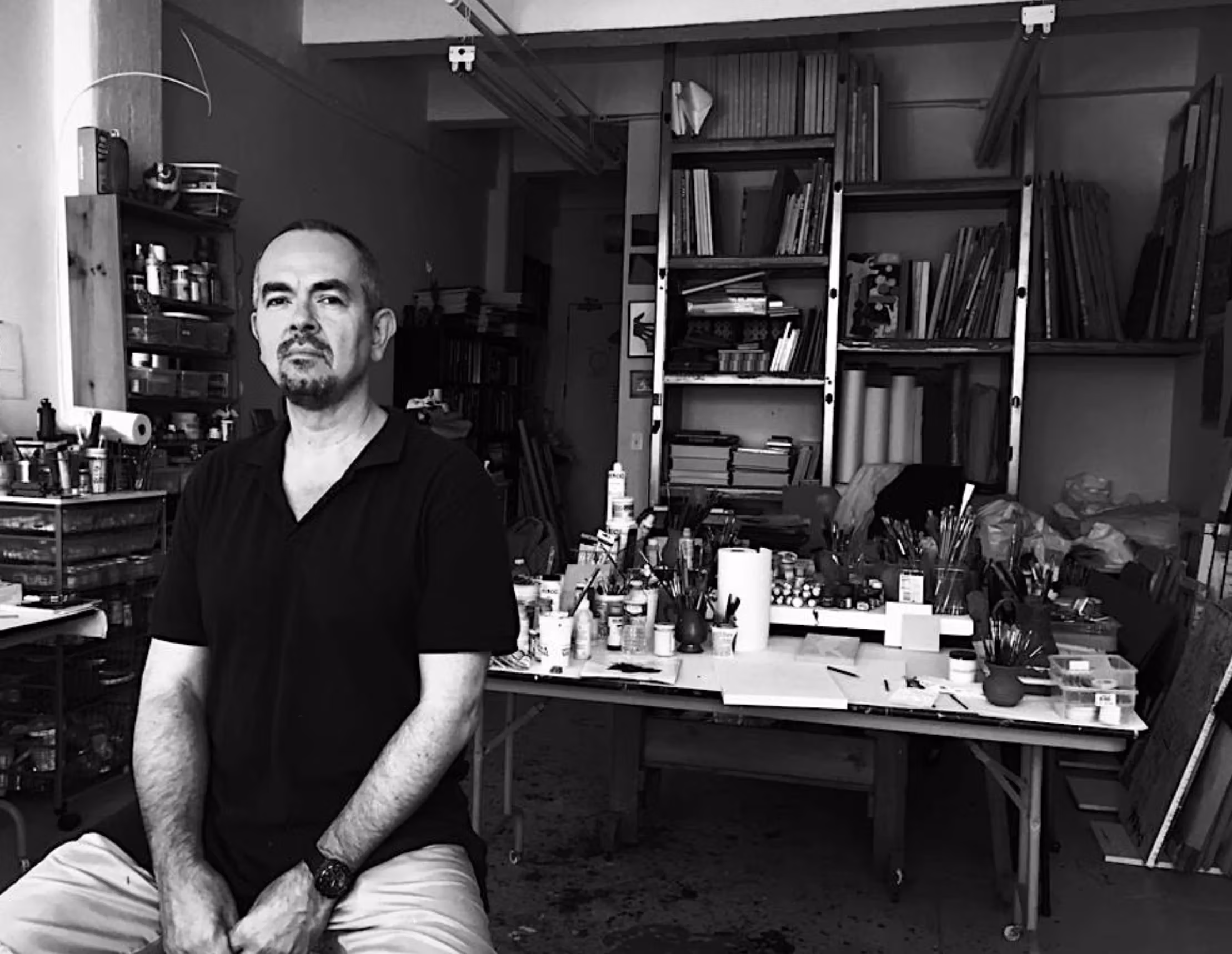 Photo of a man in a black v neck shirt, Peter Hristoff, in his studio surrounded by artwork in progress and supplies.