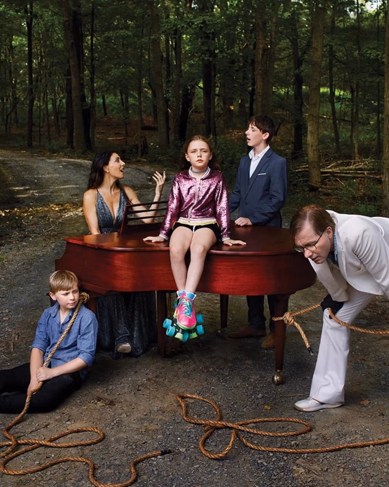 Picture of a family surrounding a grand piano in the woods. A little girl sits on top of the piano, staring at the camera. 