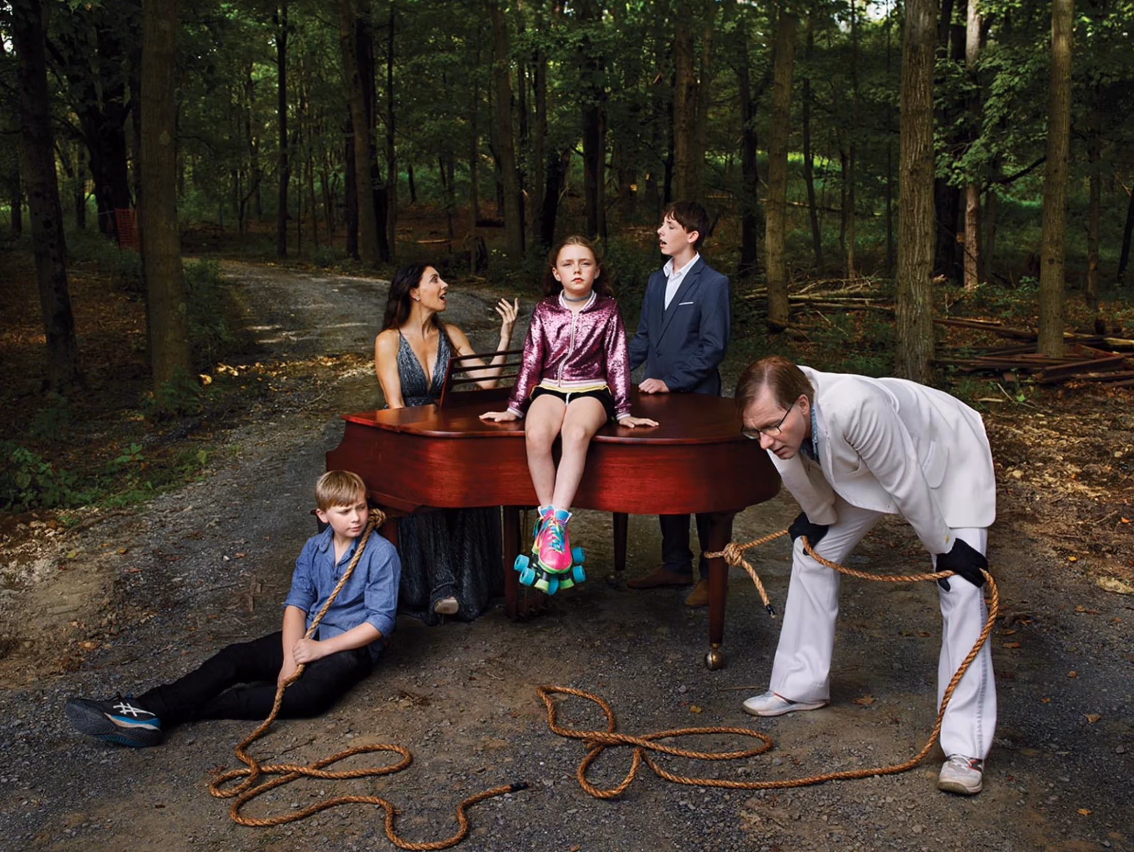 Picture of a family surrounding a grand piano in the woods. A little girl sits on top of the piano, staring at the camera. 