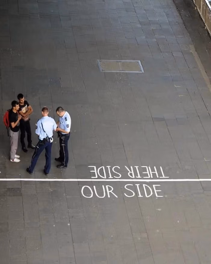 An overhead view of two police officers stopping two teen boys of color. There's a line drawn on the ground and it's said "THEIR SIDE" on the side of the line they are on and "OUR SIDE" on the other side of the line.