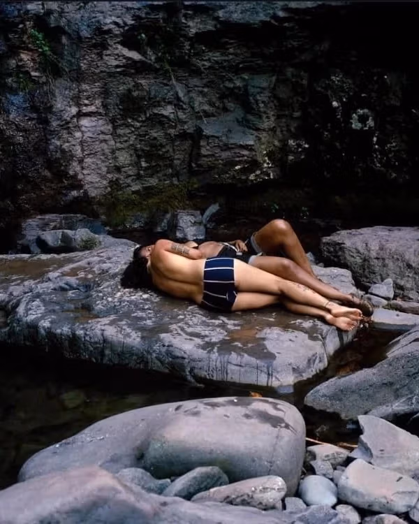 Two individuals lie in their undergarments on a rock in a calm river. There are many other rocks in the foreground.