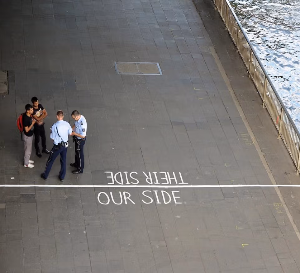 An overhead view of two police officers stopping two teen boys of color. There's a line drawn on the ground and it's said "THEIR SIDE" on the side of the line they are on and "OUR SIDE" on the other side of the line.