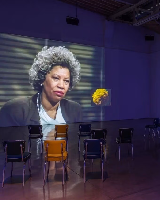 Picture of a museum installation of a large projected screen with a woman and a yellow flower. Several chairs are organized in front of the projection. 