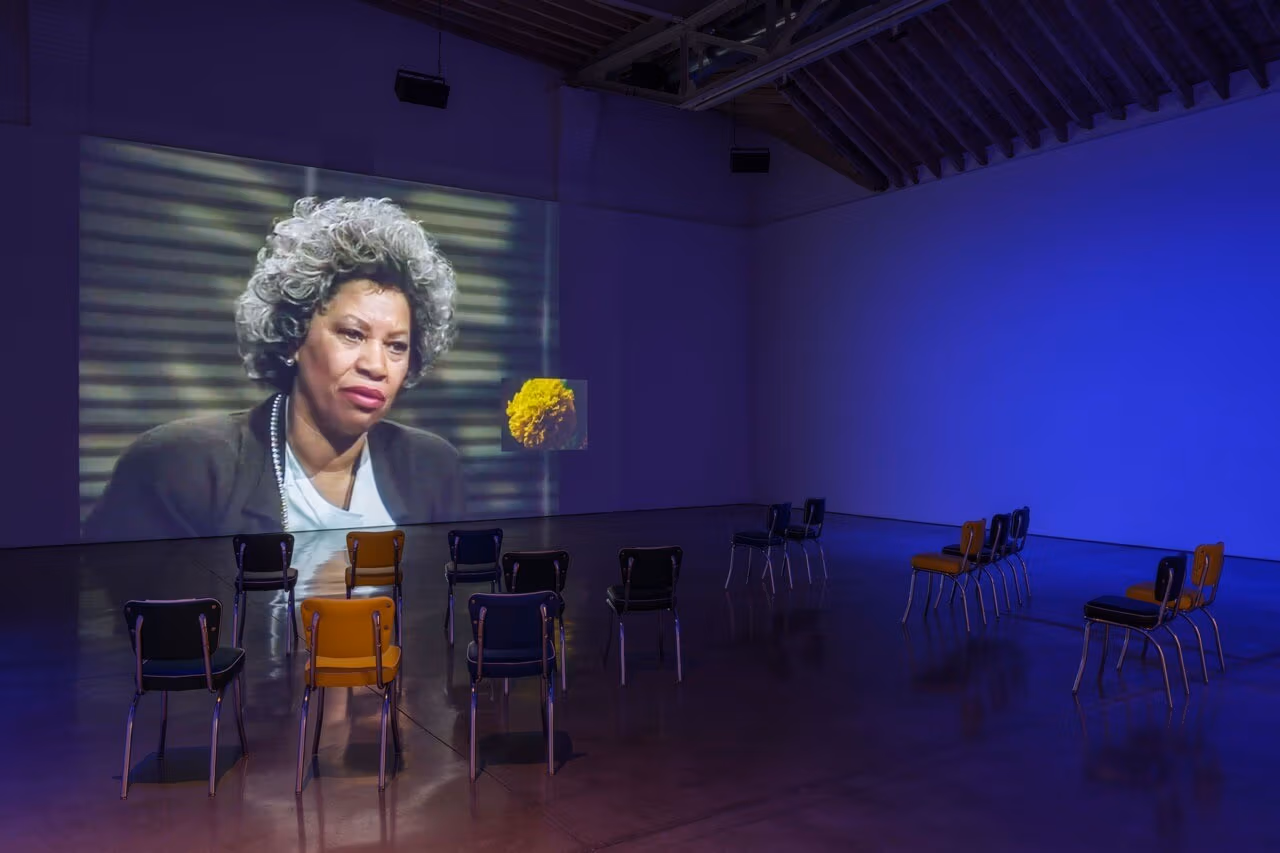 Picture of a museum installation of a large projected screen with a woman and a yellow flower. Several chairs are organized in front of the projection. 
