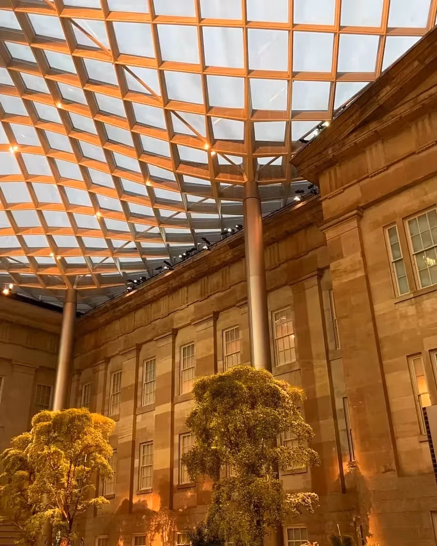 A photograph of an interior courtyard with trees, pillars, windows, and a curved glass ceiling supported by a mesh.