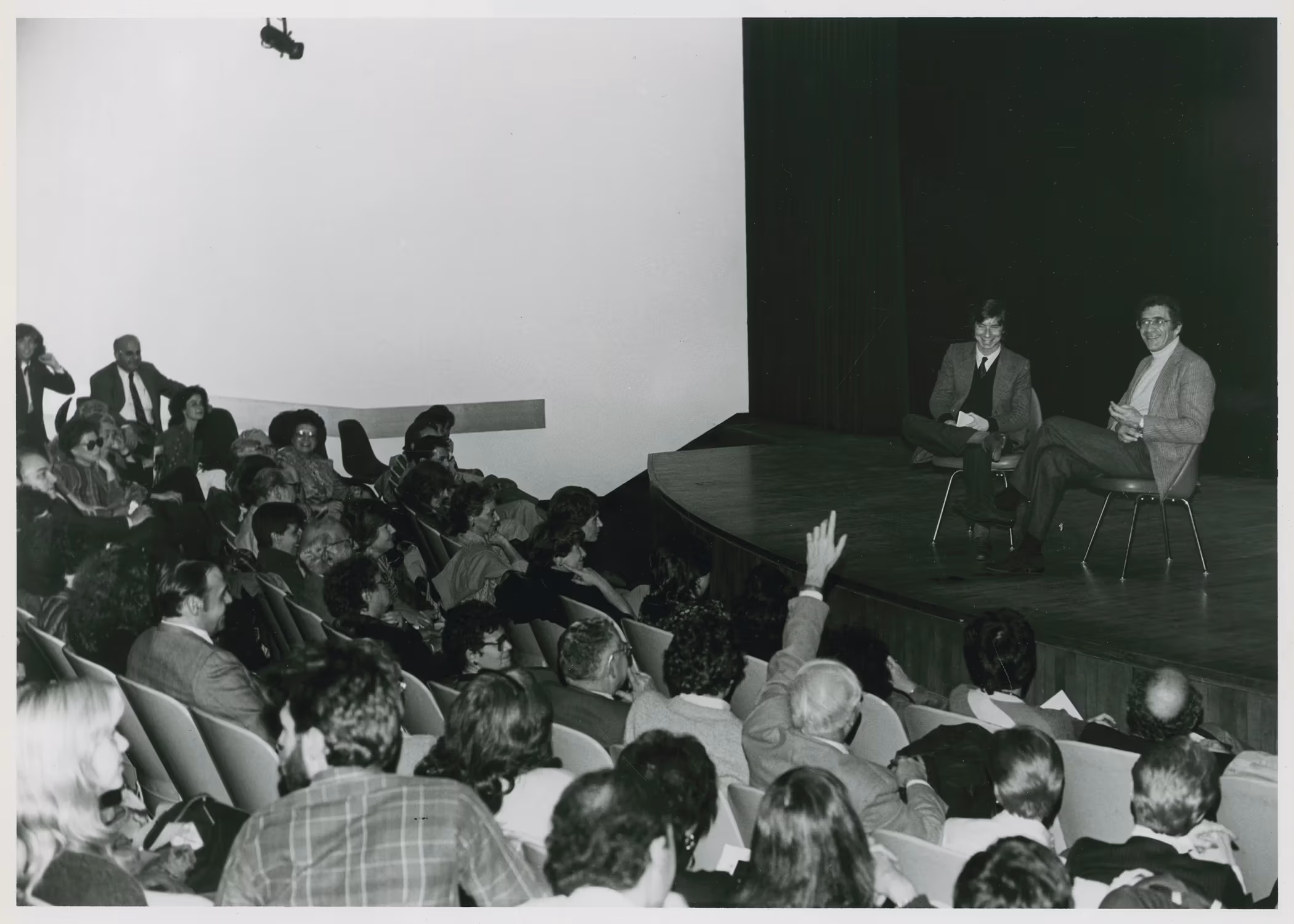 A black-and-white photo of two people sitting on an empty stage in front of a crowd. A person in the crowd has raised their hand to ask a question