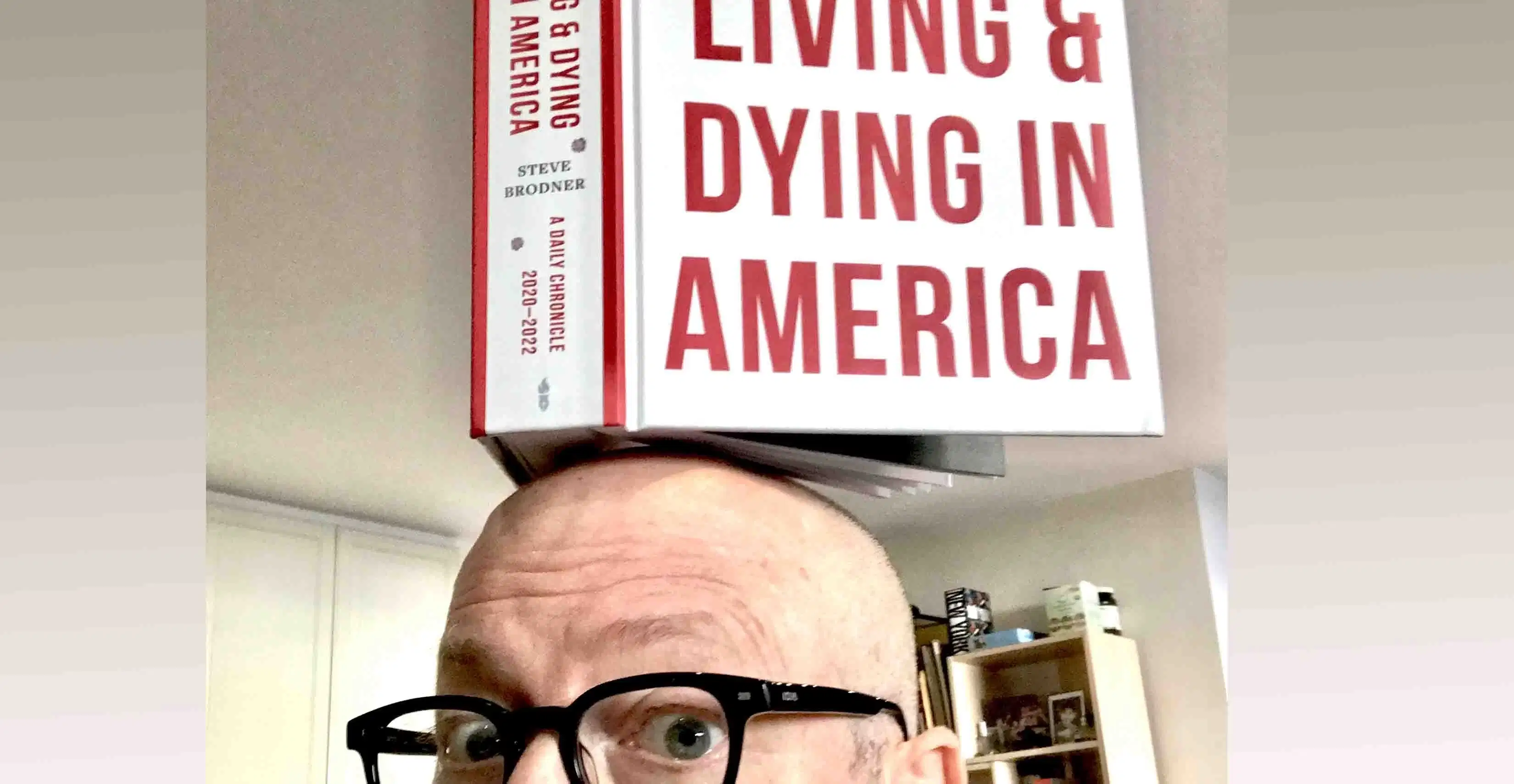 A photograph of a bald white man wearing black-rimmed glasses and balancing a book on his head.