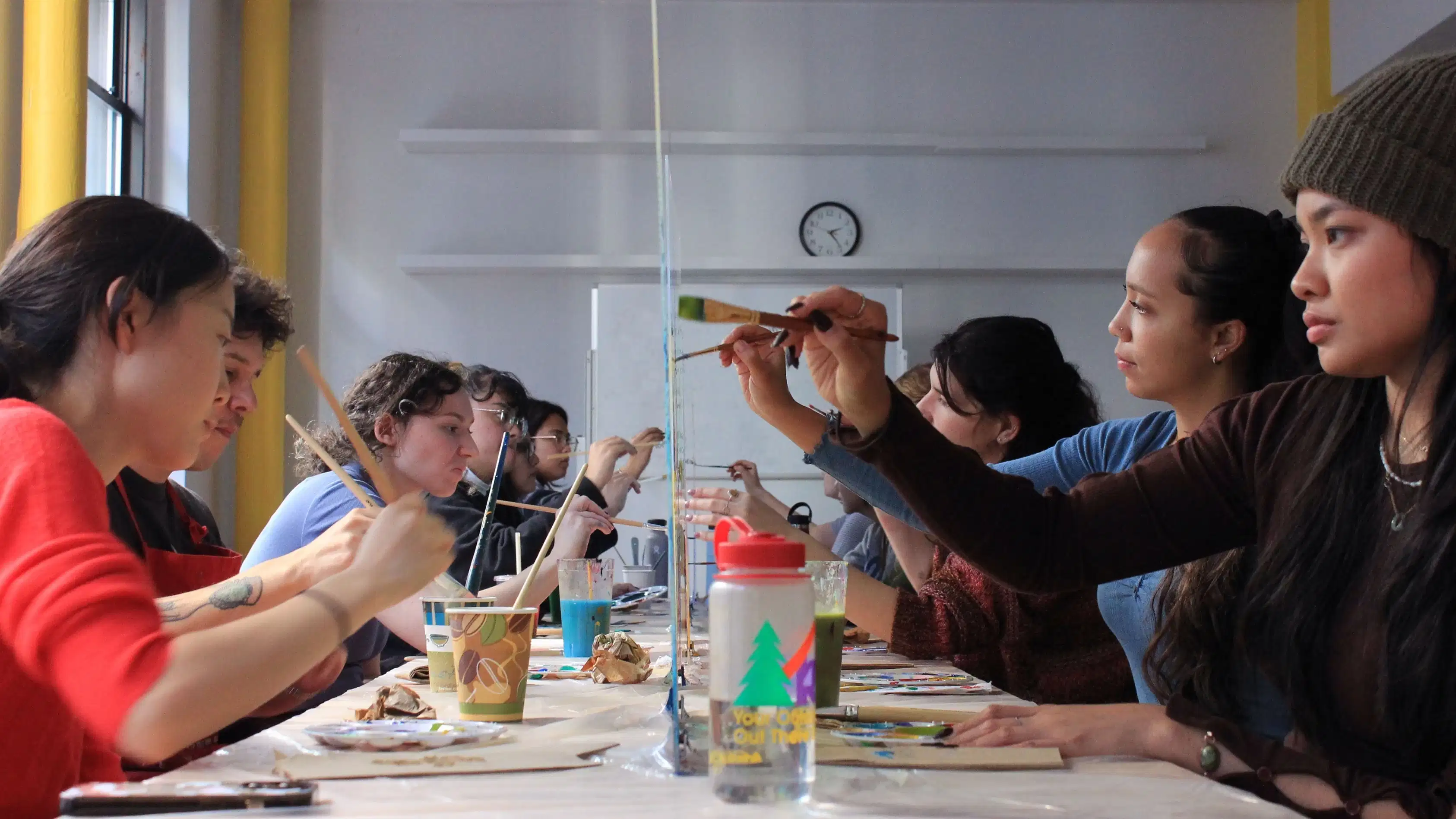 graduate students painting on either side of a plexiglass surface facing each other with paint and paintbrushes 