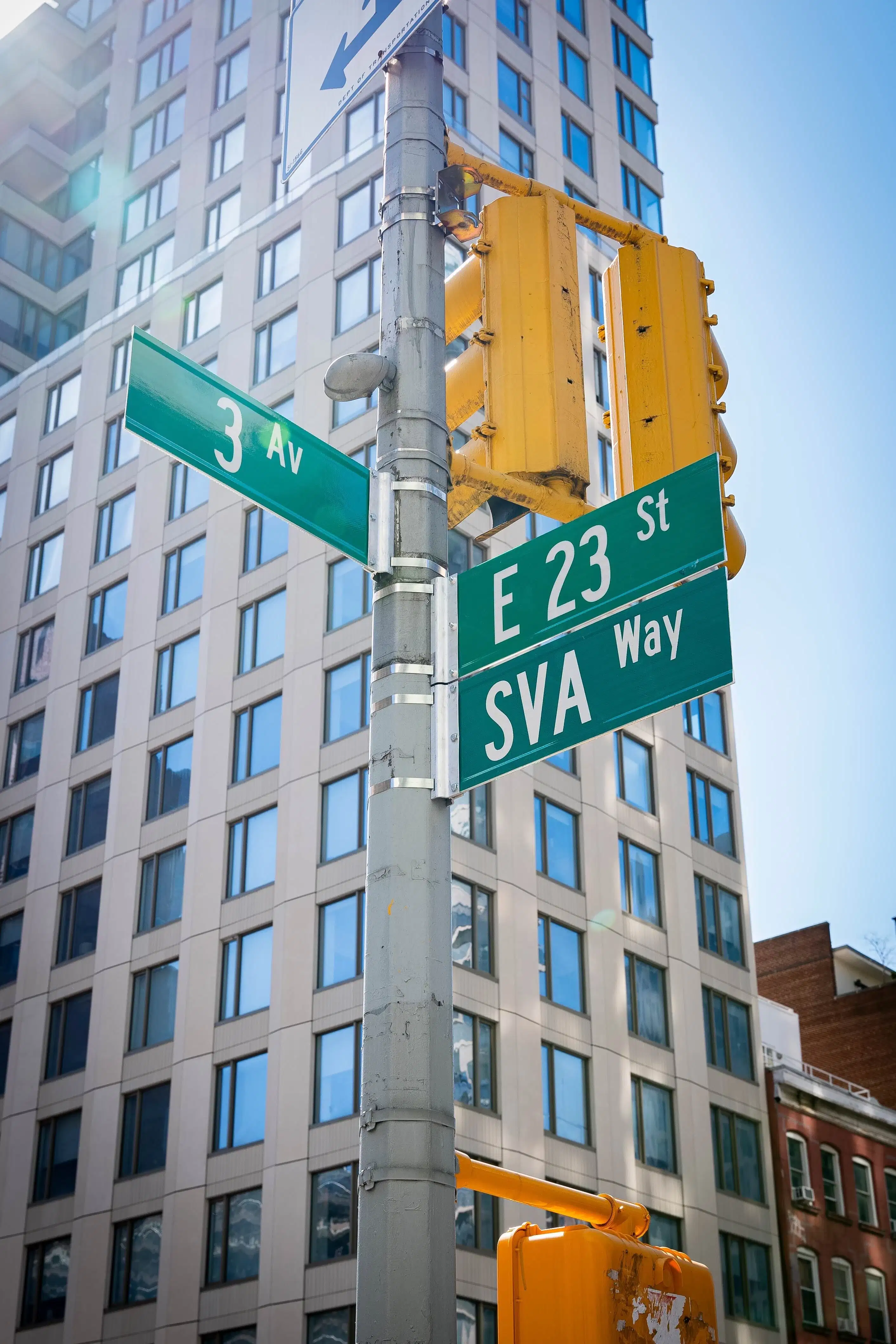 A pole with three street signs for Third Avenue, East 23rd Street, and SVA Way. A tall, modern building stands in the background. The perspective looking up at SVA Way emphasizes the honor of the co-naming.