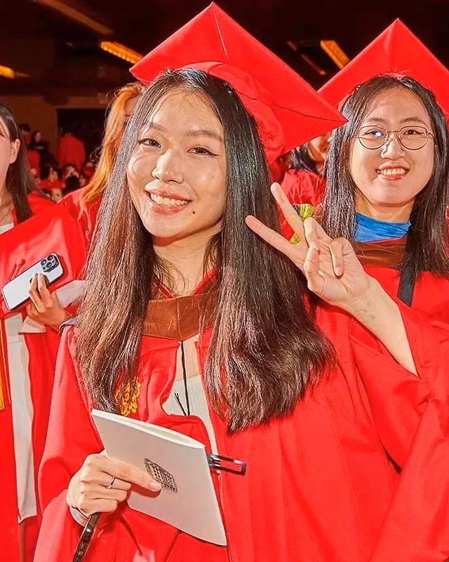 A group of students wearing red graduation gowns and caps smile and hold up peace signs during a graduation ceremony.