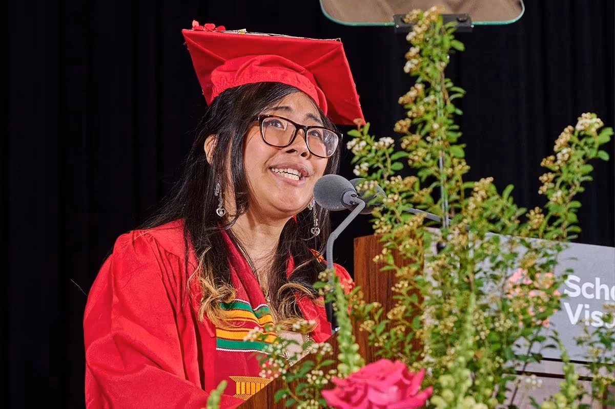 A graduate in a red cap and gown speaks passionately at a podium adorned with greenery and flowers. The background is a dark curtain, conveying a celebratory tone.
