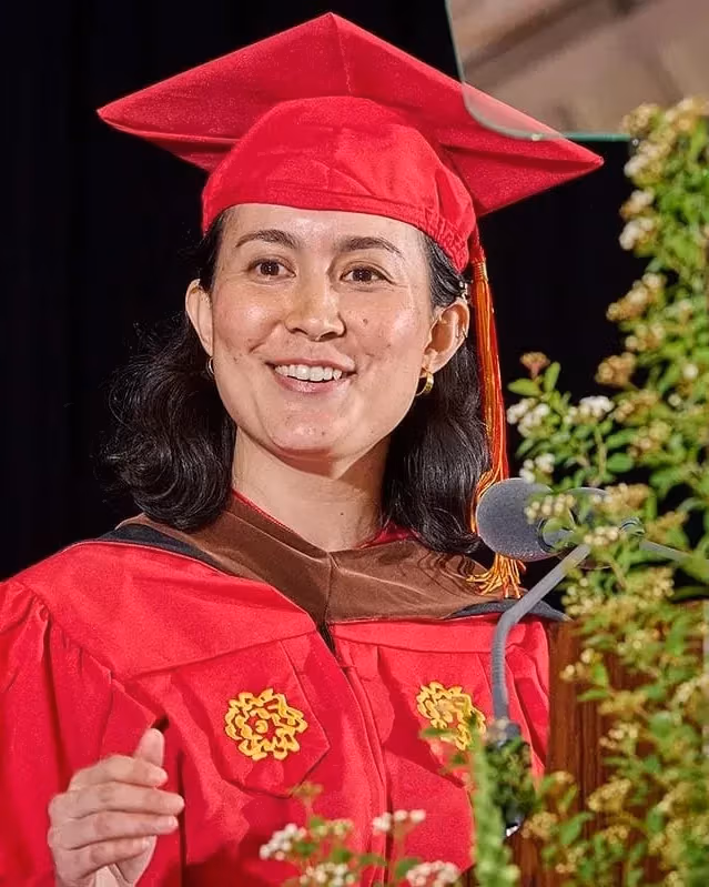 Graduate woman in red cap and gown delivers a speech at a lectern with greenery. She smiles warmly, exuding a sense of achievement and joy.