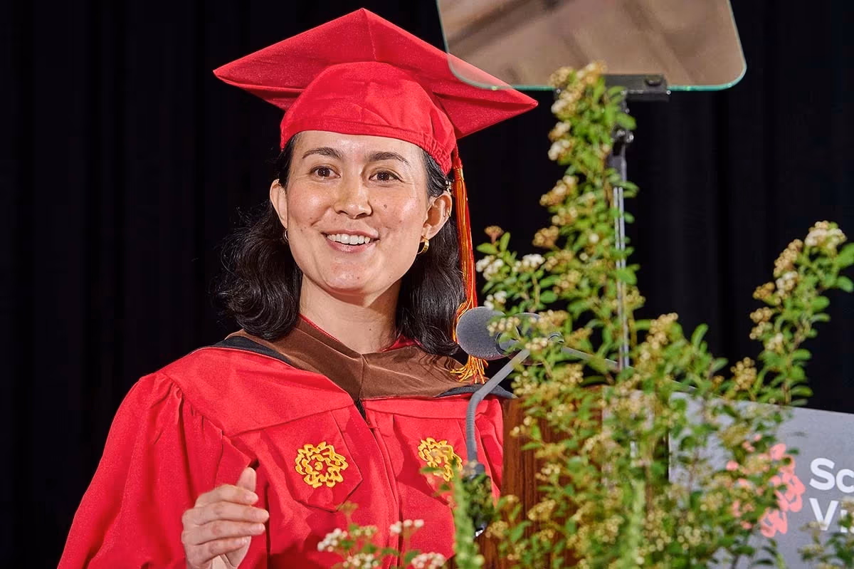 Graduate woman in red cap and gown delivers a speech at a lectern with greenery. She smiles warmly, exuding a sense of achievement and joy.