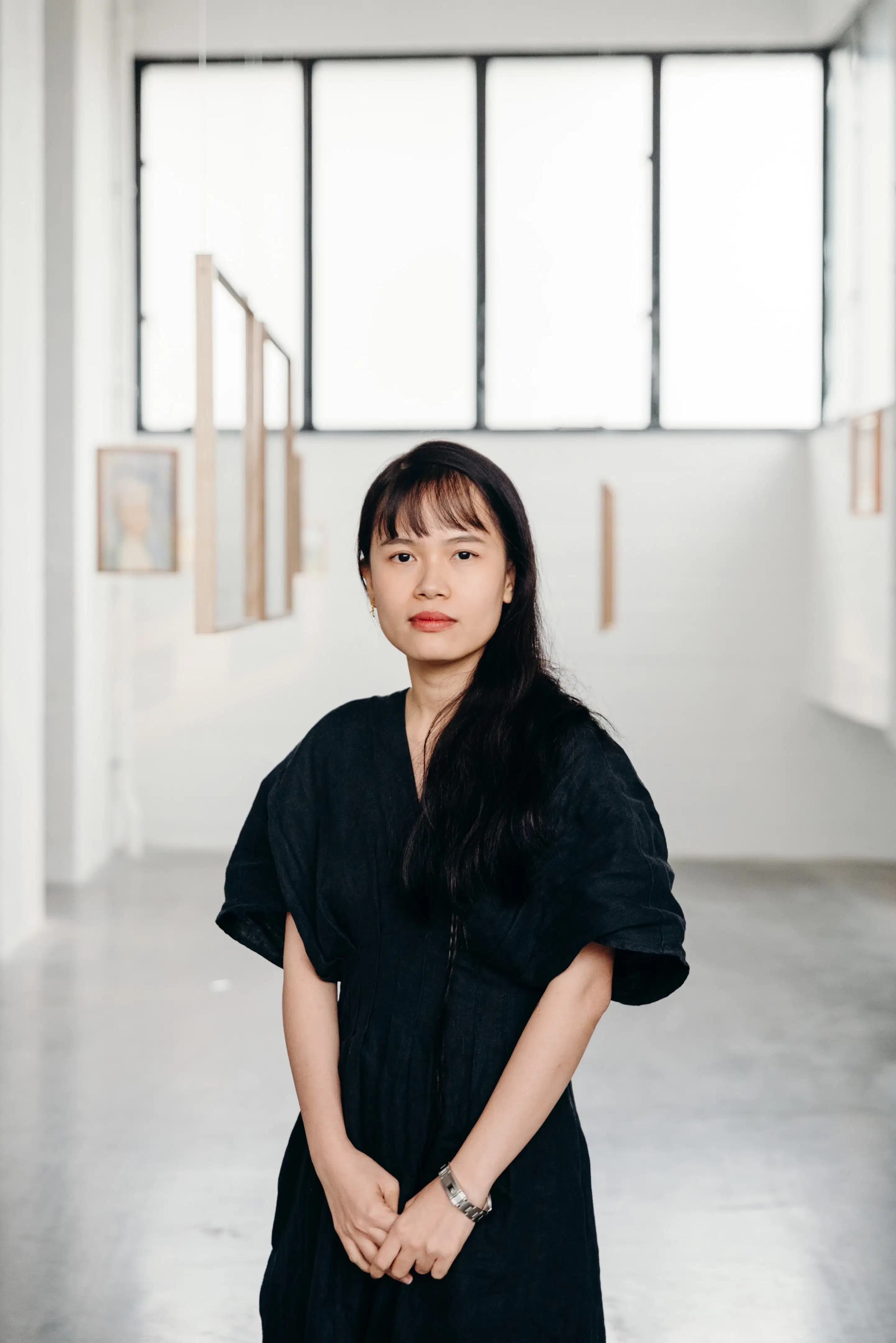 A photograph of Thao Nguyen Phan. Phan stands in the center of the photo wearing a black dress. In the background the viewer can see some of Phan's artworks hanging in a gallery.