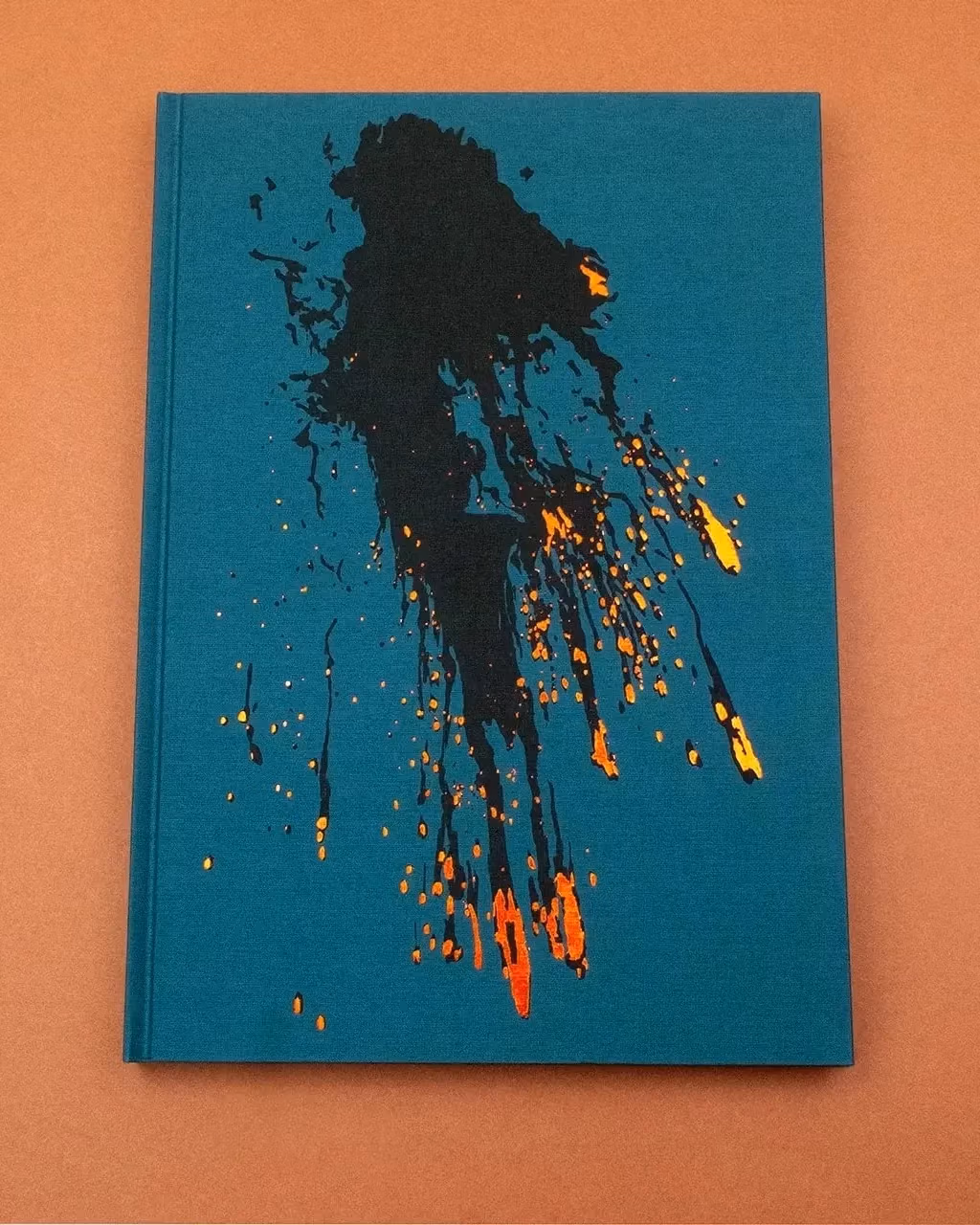 A teal book cover on a muted orange background. On the cover there's a splash of black ink with bright specks of orange, yellow, and red on the tails of the ink to make the image resemble the aftermath of a mid-air explosion.