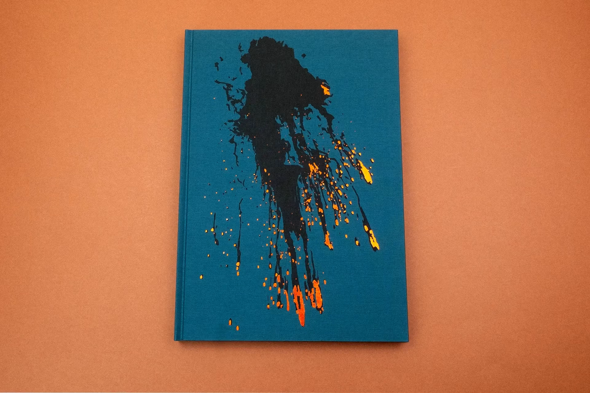 A teal book cover on a muted orange background. On the cover there's a splash of black ink with bright specks of orange, yellow, and red on the tails of the ink to make the image resemble the aftermath of a mid-air explosion.