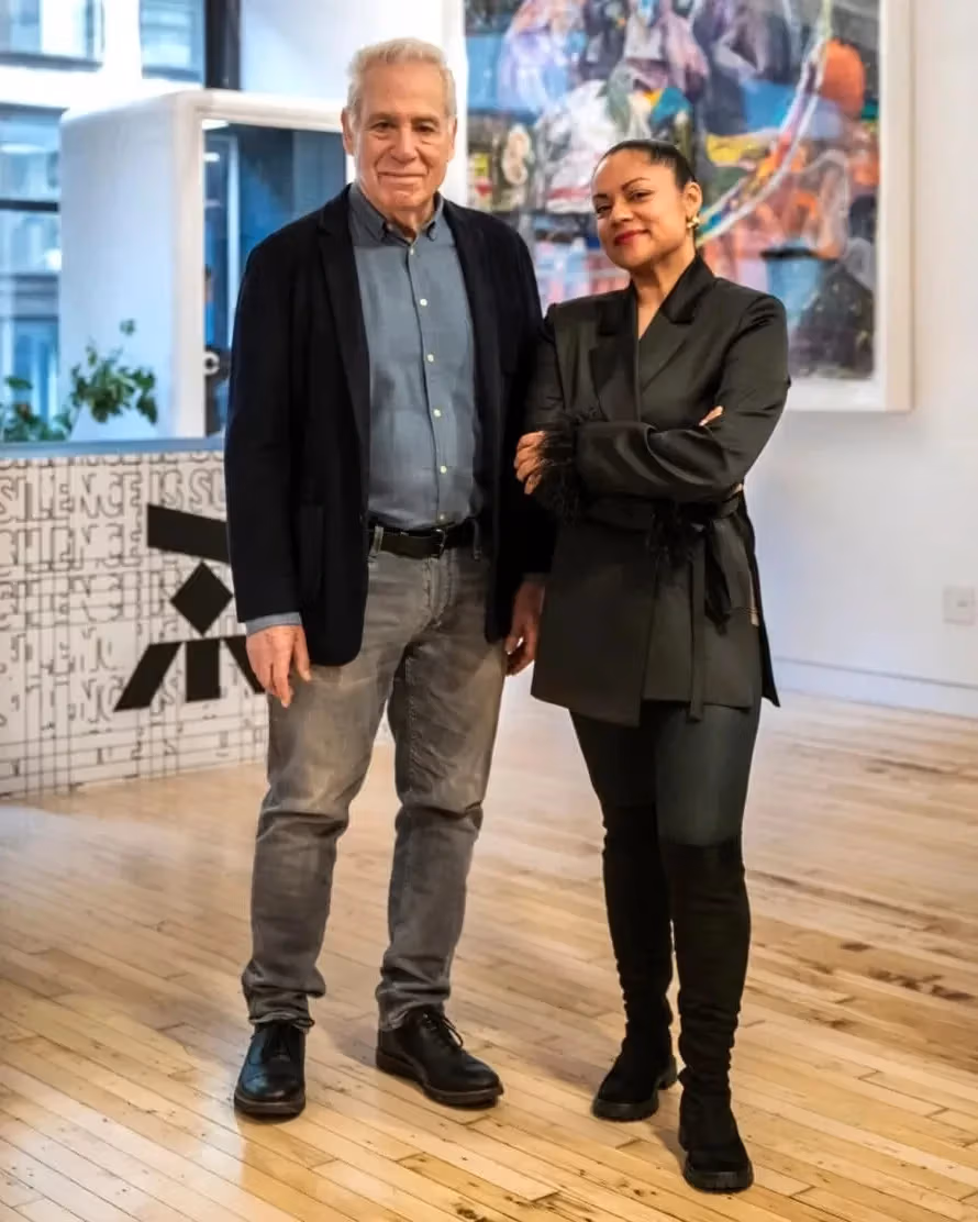 Two people stand side by side in a modern, well-lit room with wooden floors, artwork on the wall, and a decorative partition in the background.