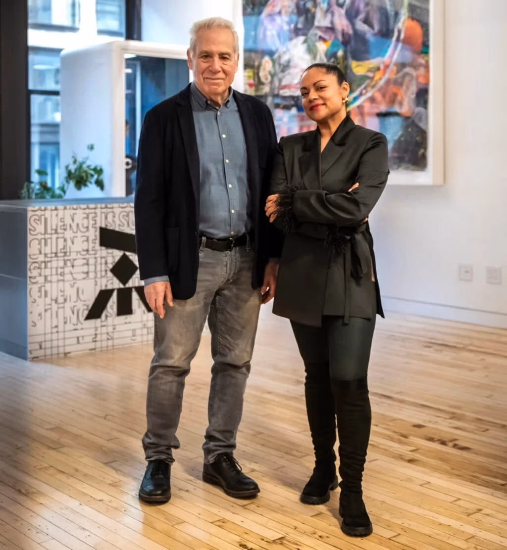 Two people stand side by side in a modern, well-lit room with wooden floors, artwork on the wall, and a decorative partition in the background.