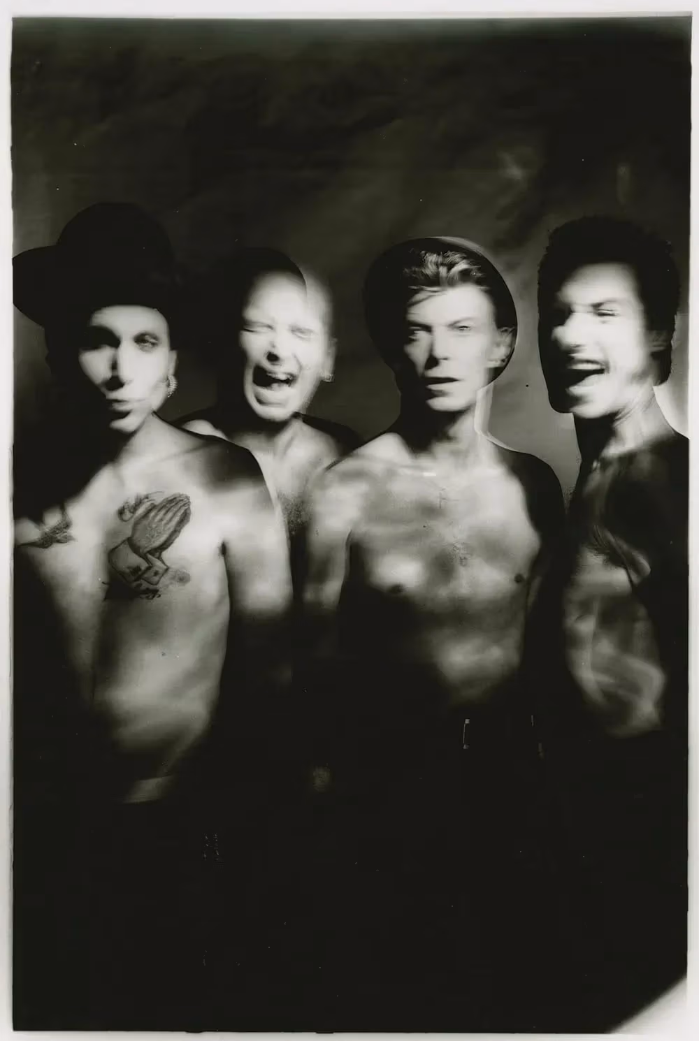 Four shirtless people with blurred faces standing together; one has a hat and a praying hands tattoo, another has slicked-back hair, all are in a dark, smoky setting.